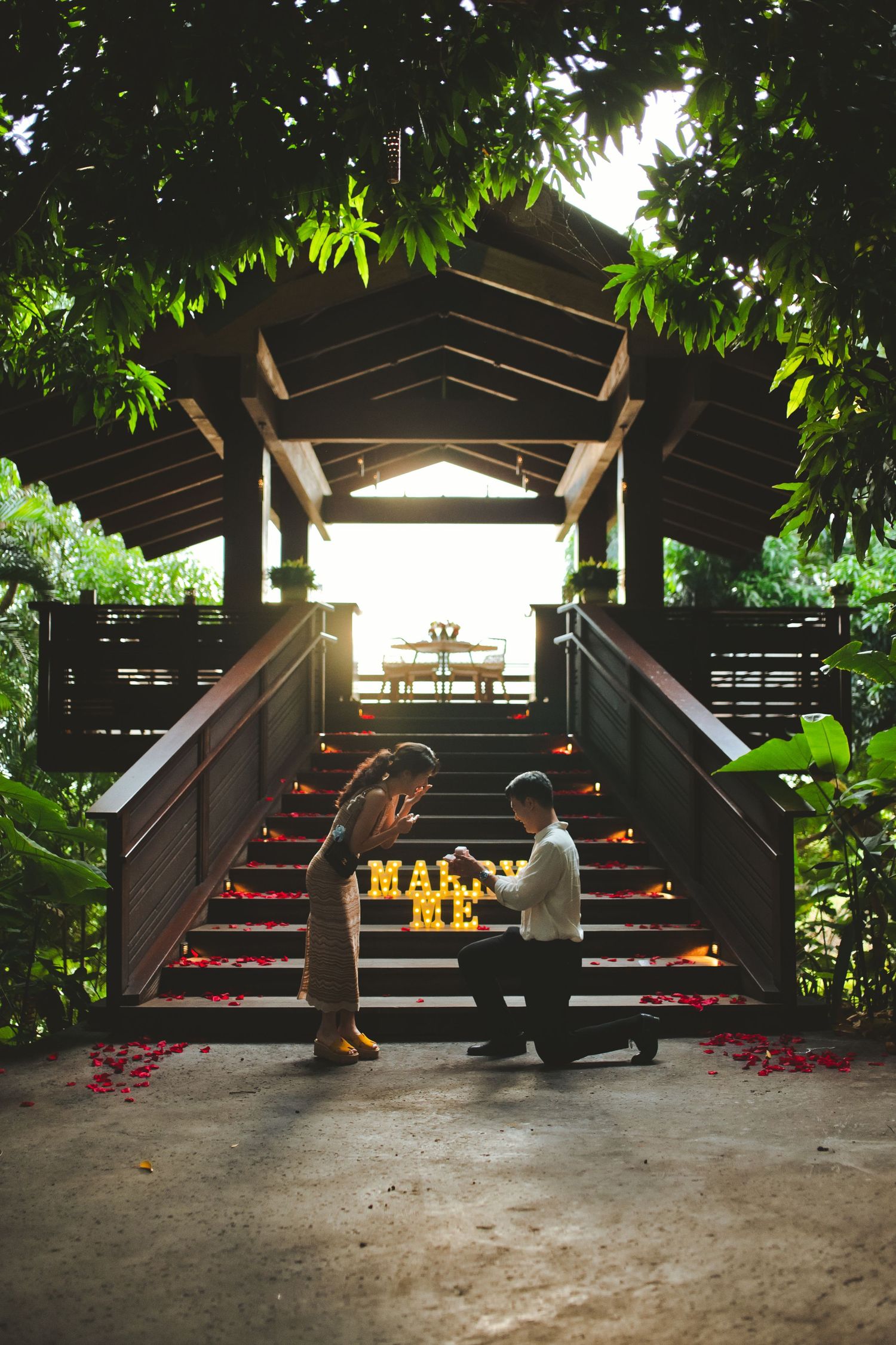 Man proposes to woman on decorated tropical stairway with candles and rose petals at sunset.