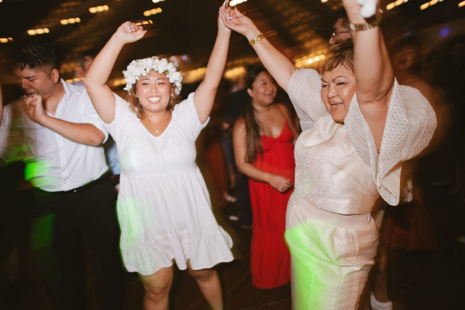 Joyful wedding guests dancing at reception, woman wearing floral crown with hands raised.