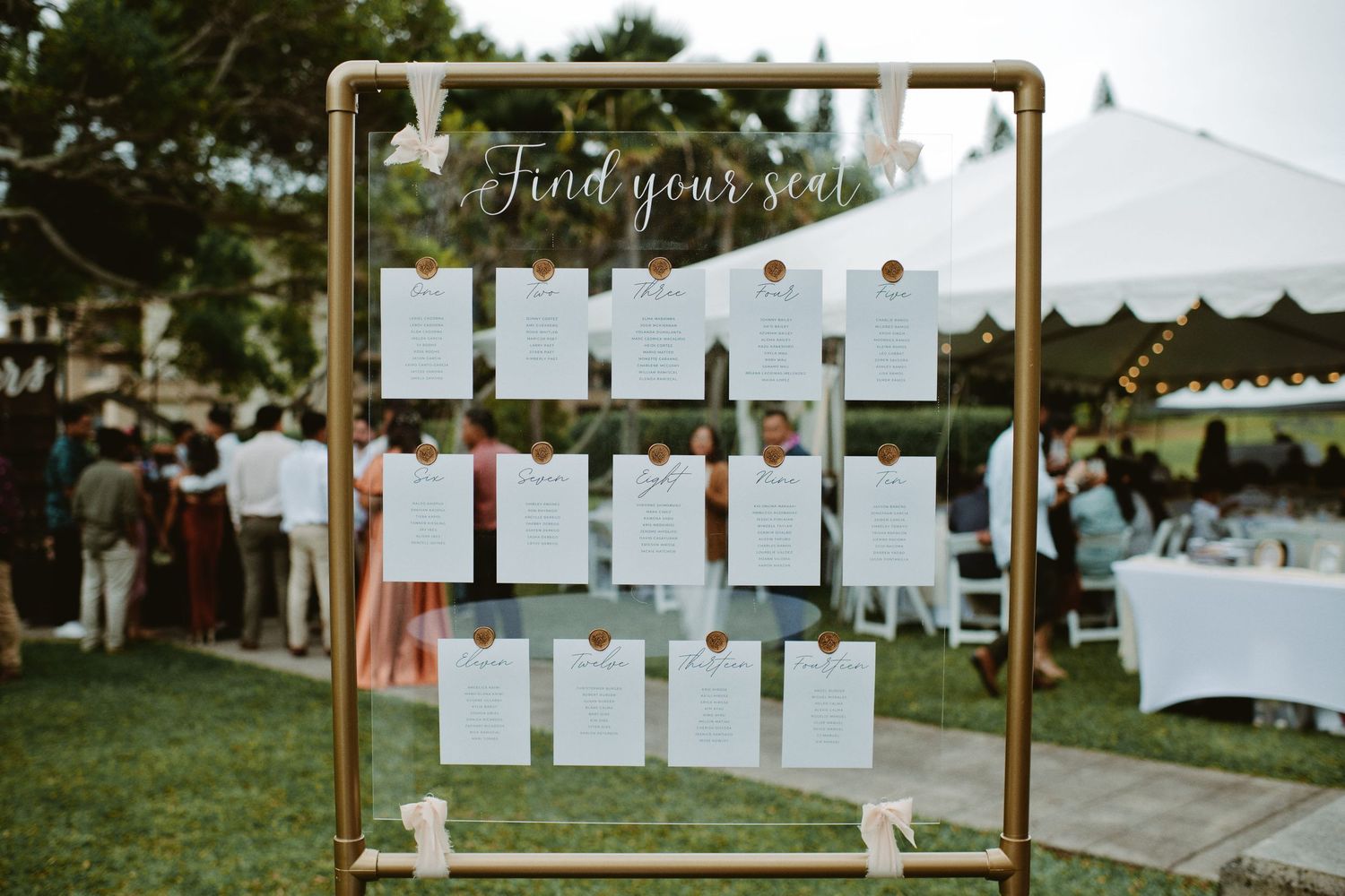 Gold pipe frame wedding seating chart with white cards on a lawn, reading Find Your Seat at an outdoor reception.