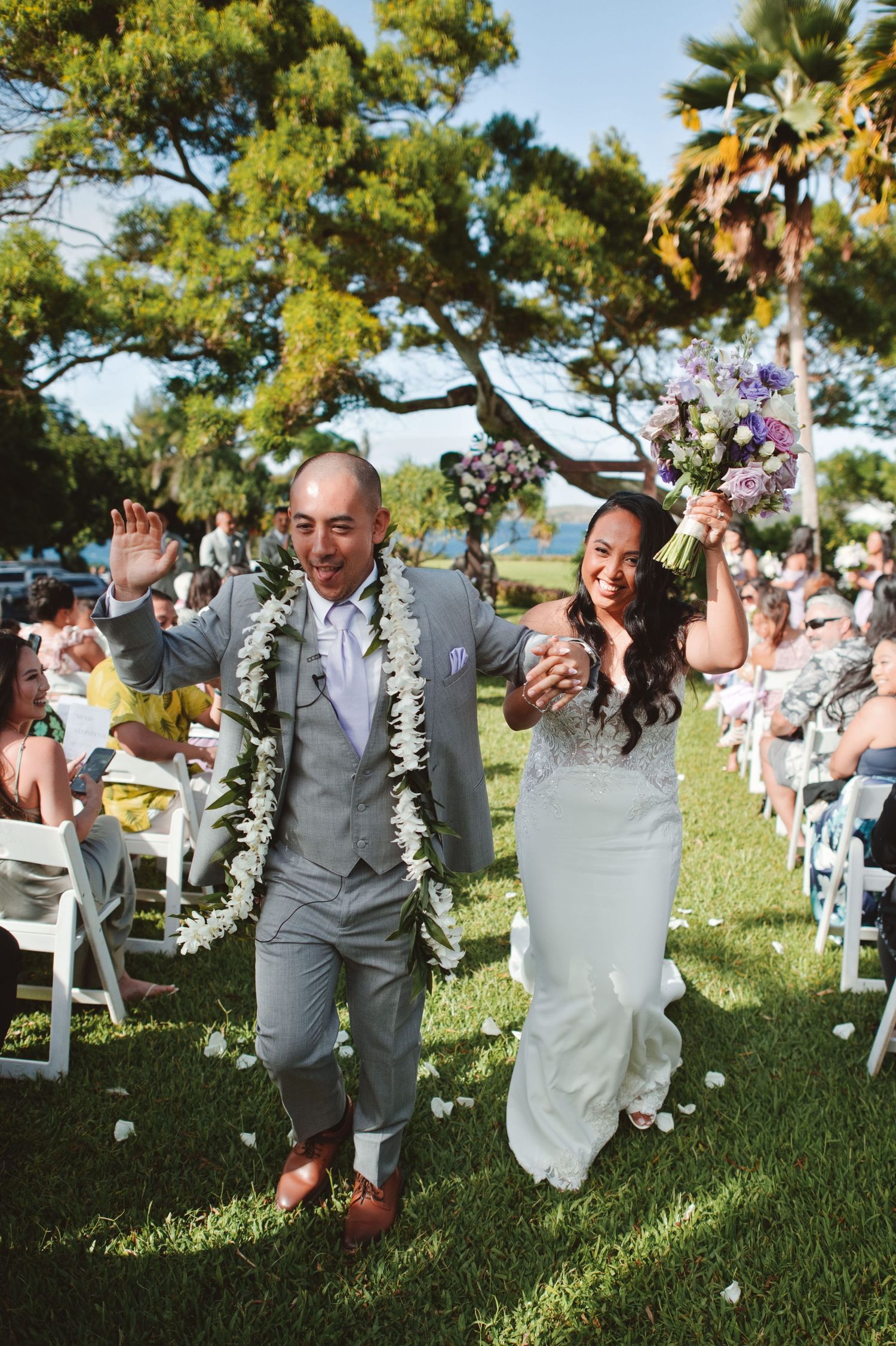 Joyful newlyweds walk down the aisle at an outdoor Hawaiian wedding ceremony, groom in gray suit with lei.