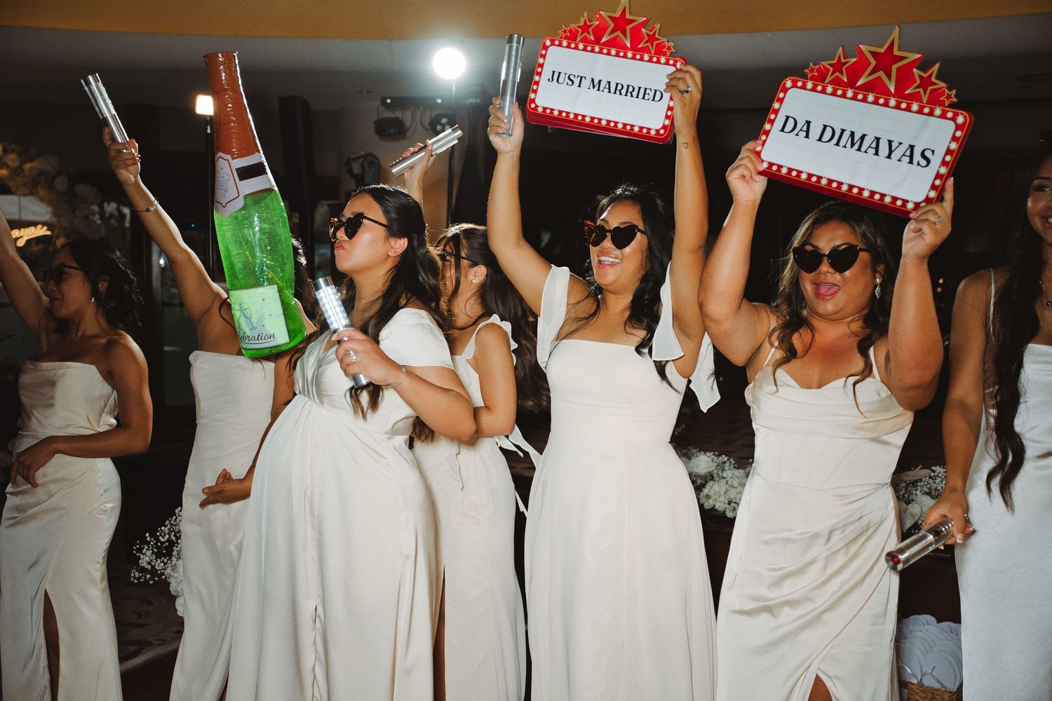 Women in white dresses celebrate at a wedding reception holding signs reading Just Married and Da Dimayas.