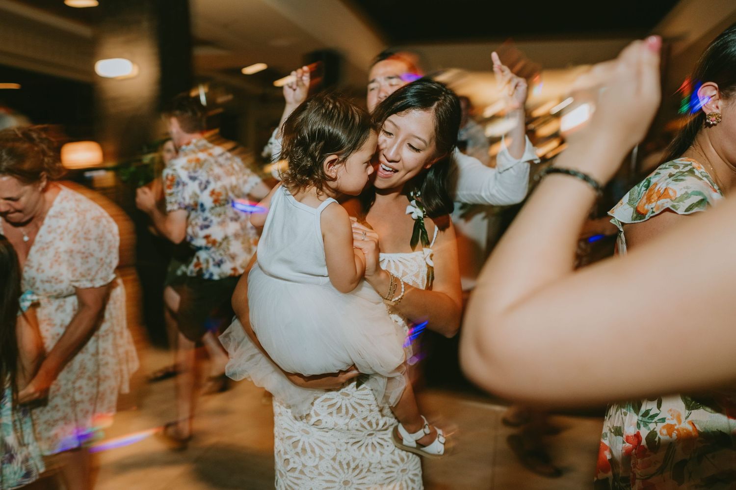 Woman in lace dress dancing and kissing a toddler at a lively wedding reception.