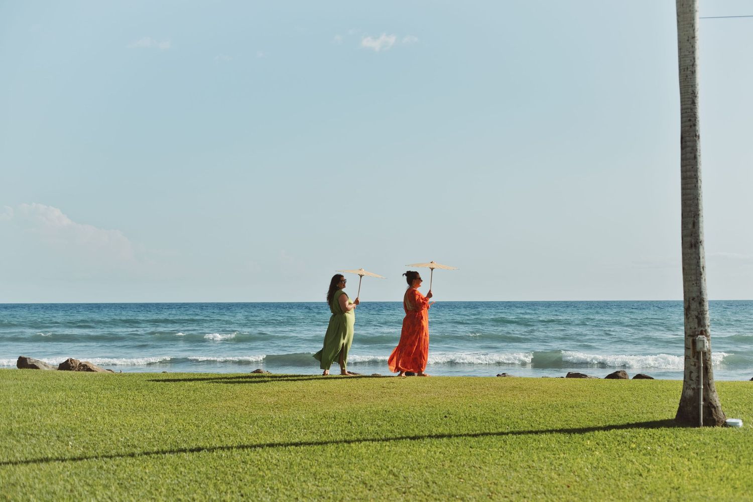 Two women in colorful maxi dresses perform a hula dance on green lawn by the ocean in Hawaii.