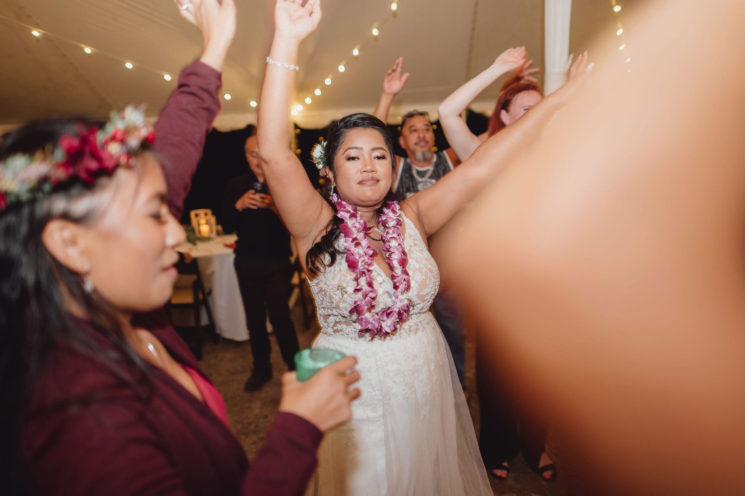 Bride in white gown and floral lei dances joyfully at wedding reception under string lights.
