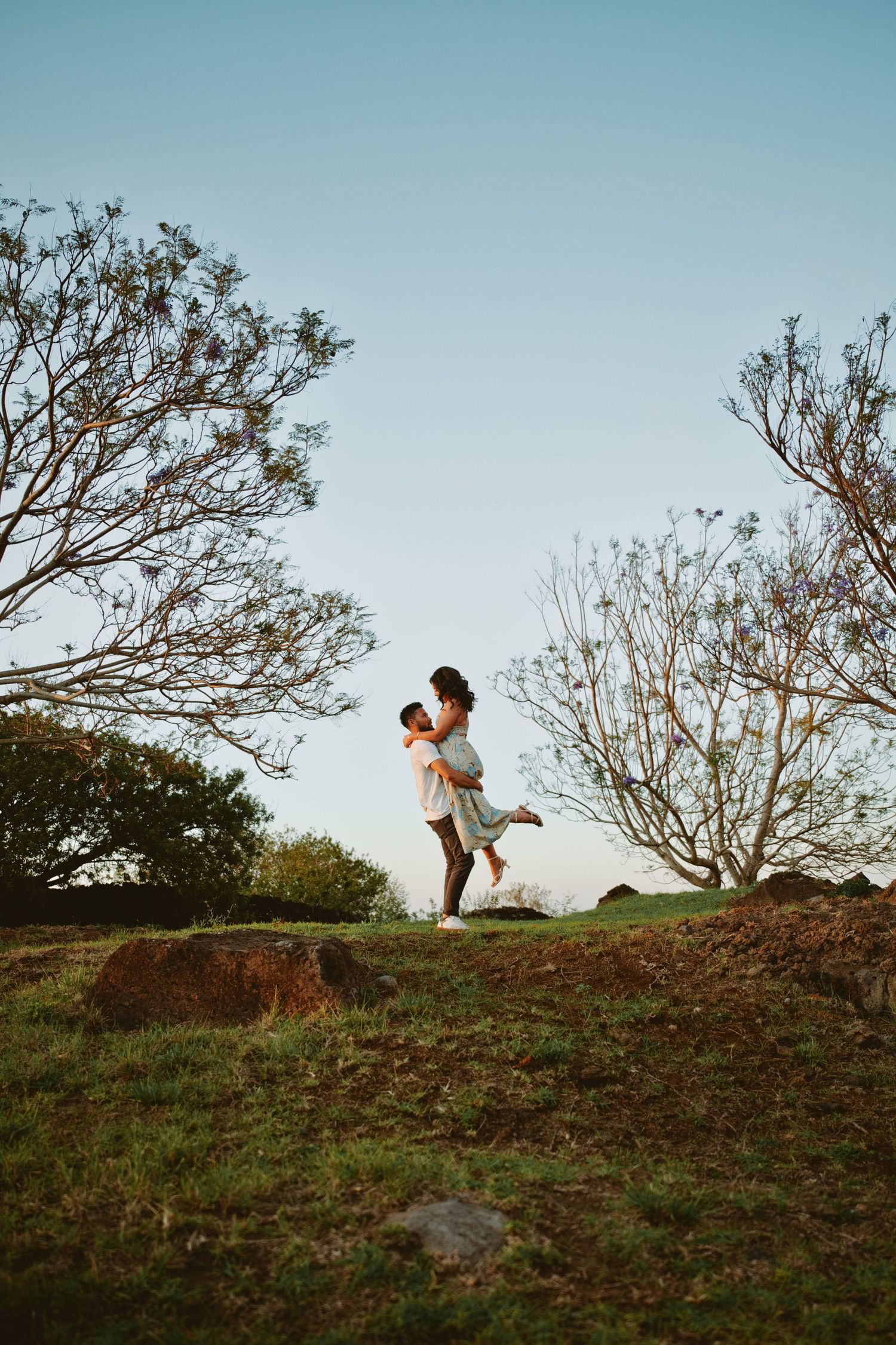 Man lifting woman in his arms on a grassy hilltop with trees at sunset.