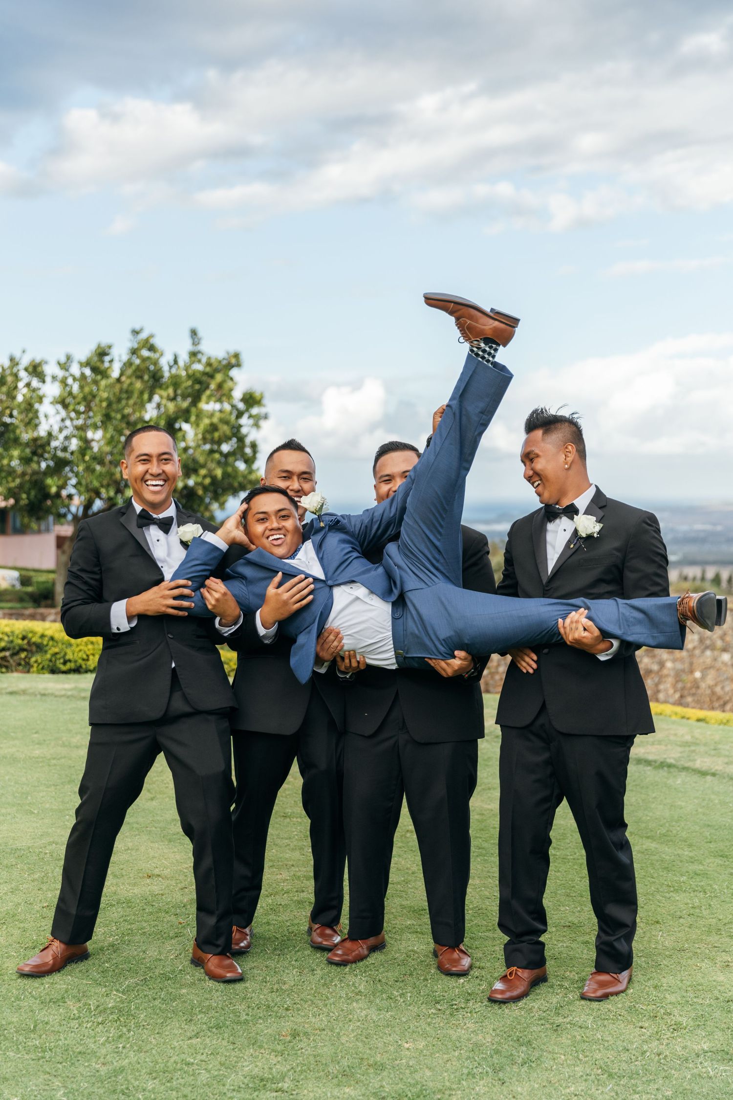 Groomsmen in black tuxedos playfully lift and hold one of their group members horizontally.