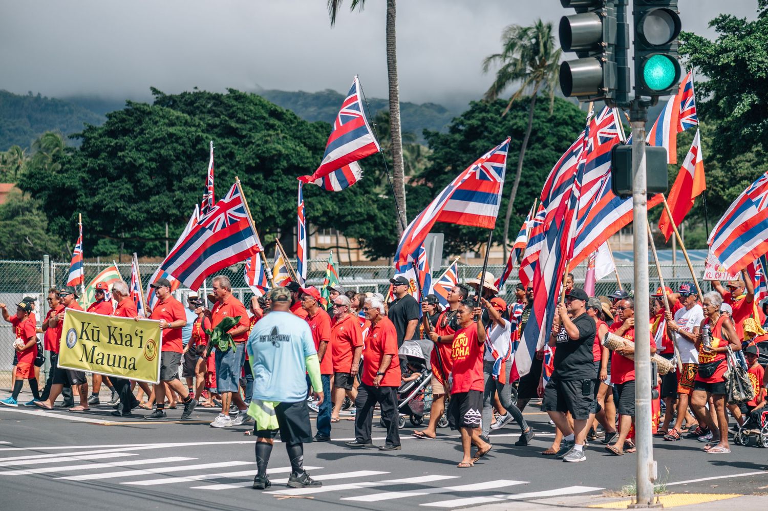 Large crowd carrying red and Hawaiian flags marches down a street with mountains in the background.