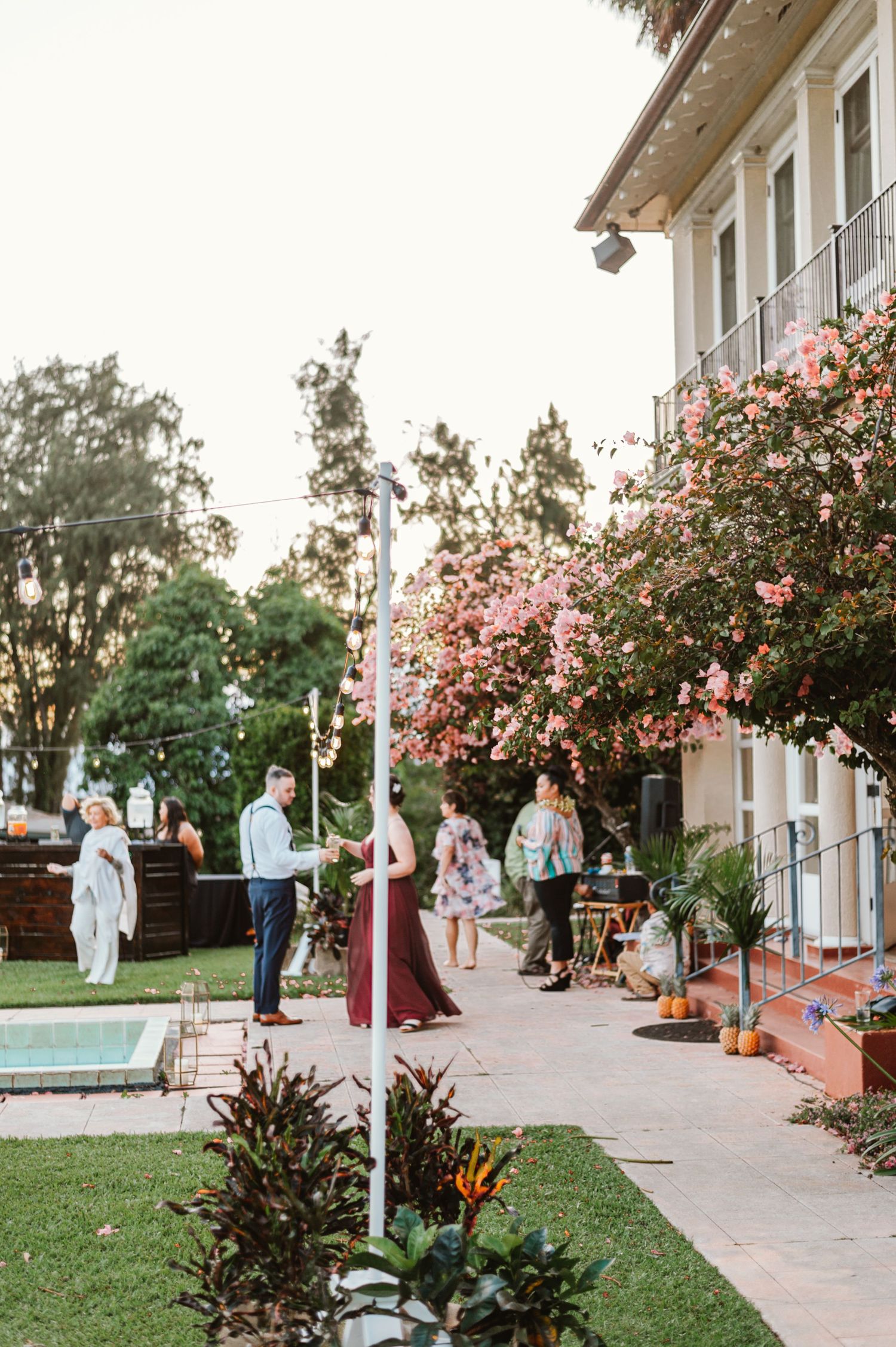 Wedding guests mingle on an elegant patio with pink flowering trees and a pool in the background.