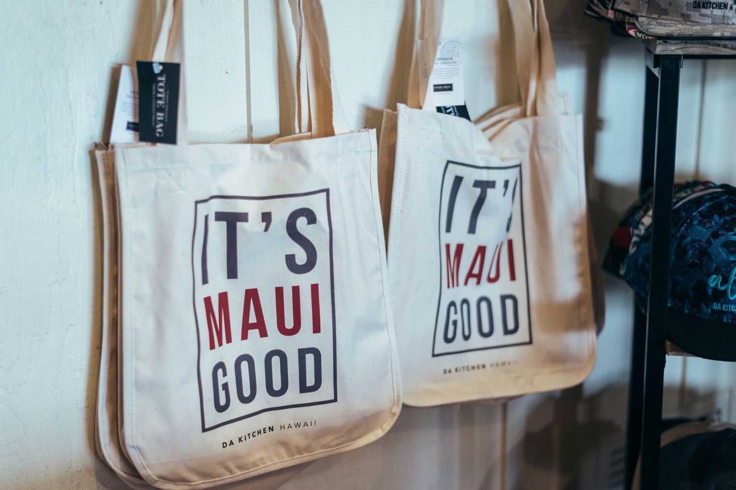 Canvas tote bags with 'It's Maui Good' text displayed in retail setting.