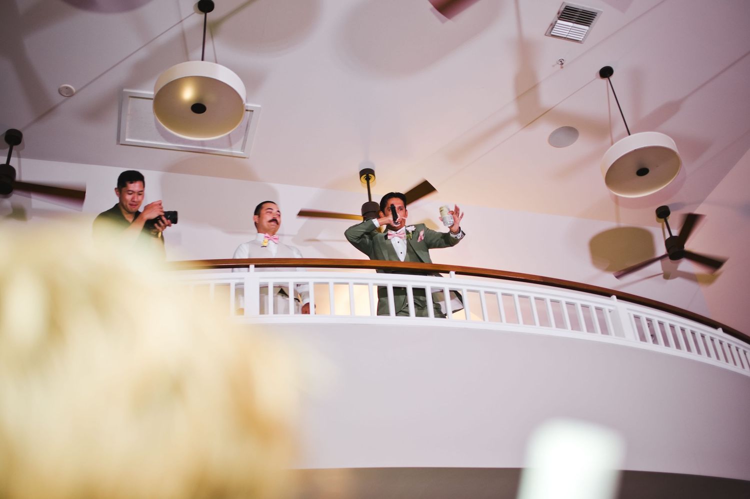 Three men in suits celebrate on a white balcony, one raising his arms joyfully at an elegant indoor venue.