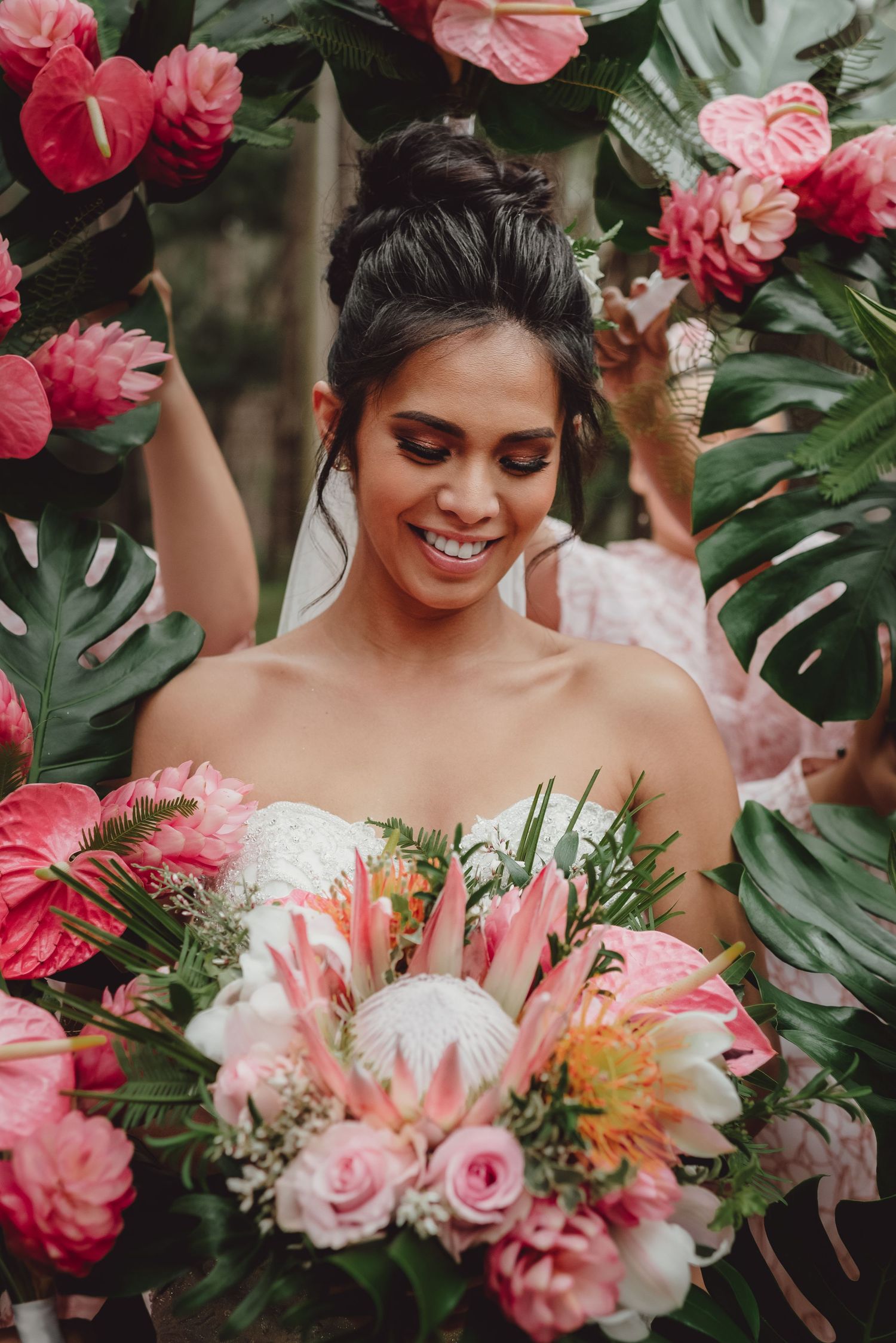 Smiling bride holding tropical pink bouquet with king protea, framed by lush monstera leaves and anthurium flowers.