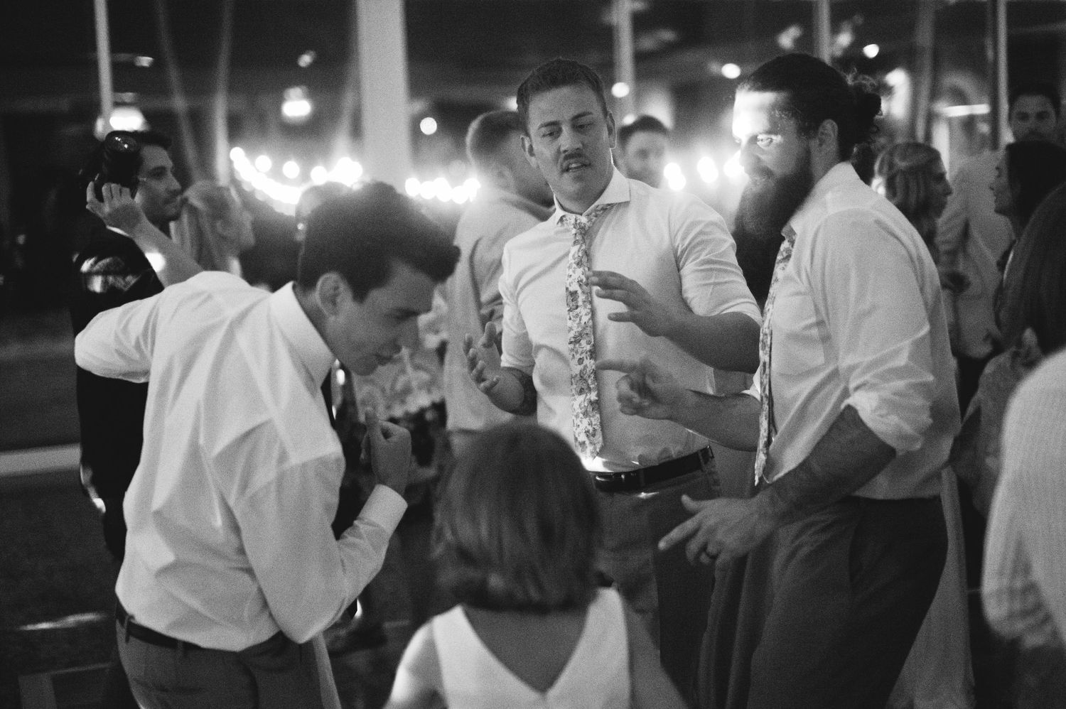 Black and white photo of men in dress shirts socializing and dancing at a wedding reception.