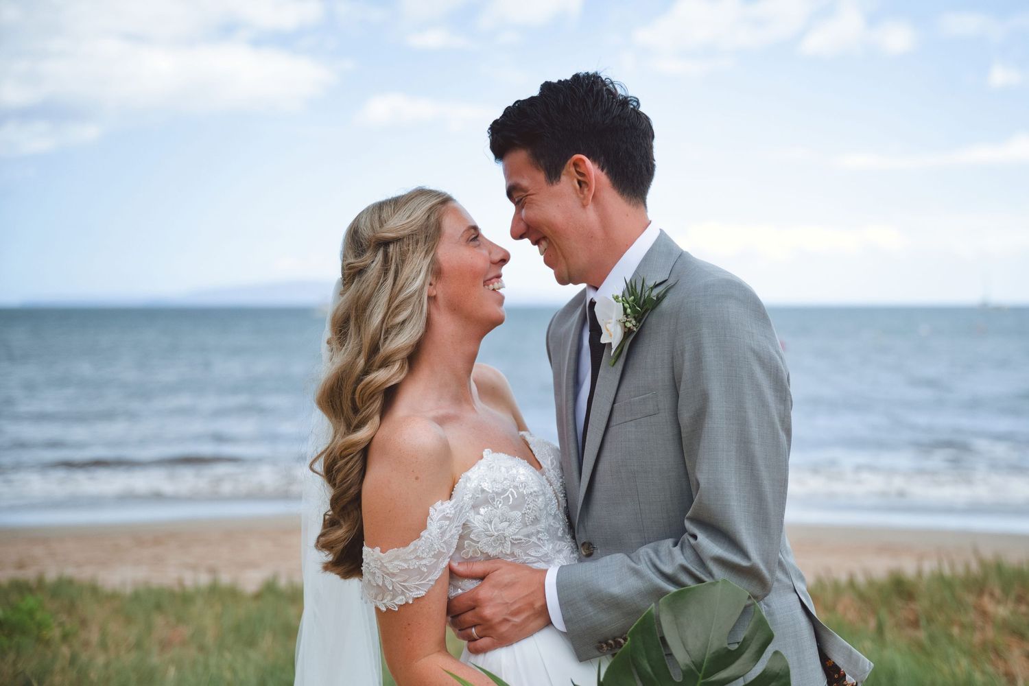 Bride and groom share a romantic moment on a beach, with ocean waves and blue sky in the background.