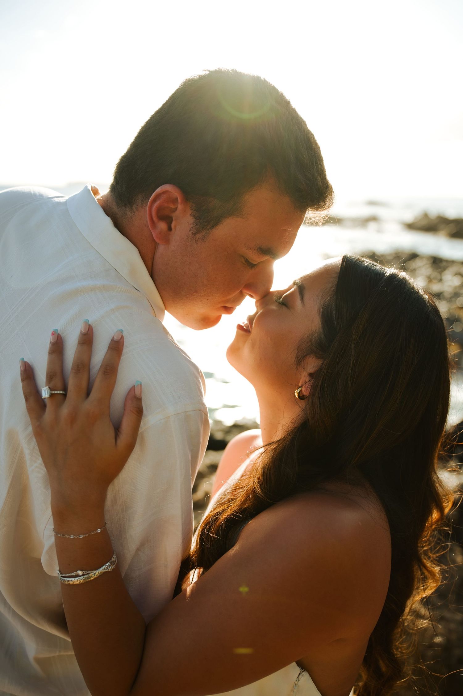 Couple sharing a kiss backlit by golden sunlight near rocky shoreline at sunset.