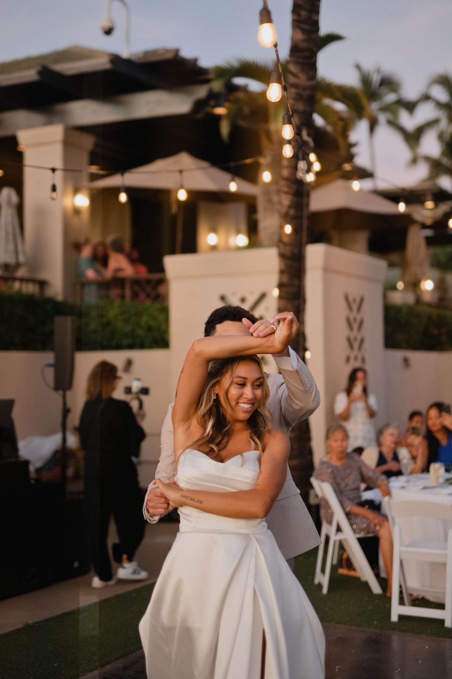 Bride and groom share their first dance outdoors at a tropical wedding reception with string lights and guests seated nearby.