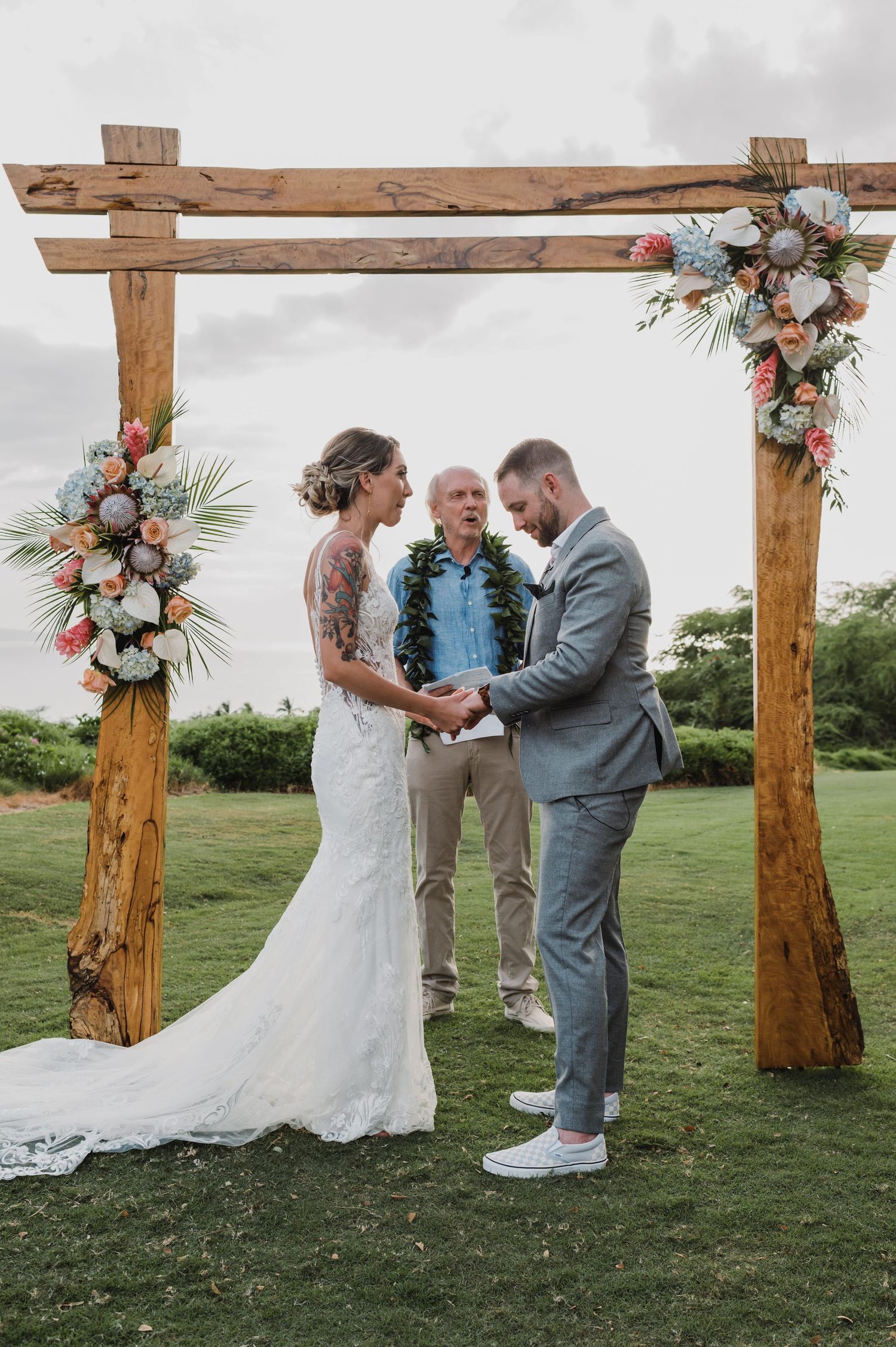 Couple exchanges vows under rustic wooden arch adorned with tropical flowers at an outdoor Hawaii wedding ceremony.