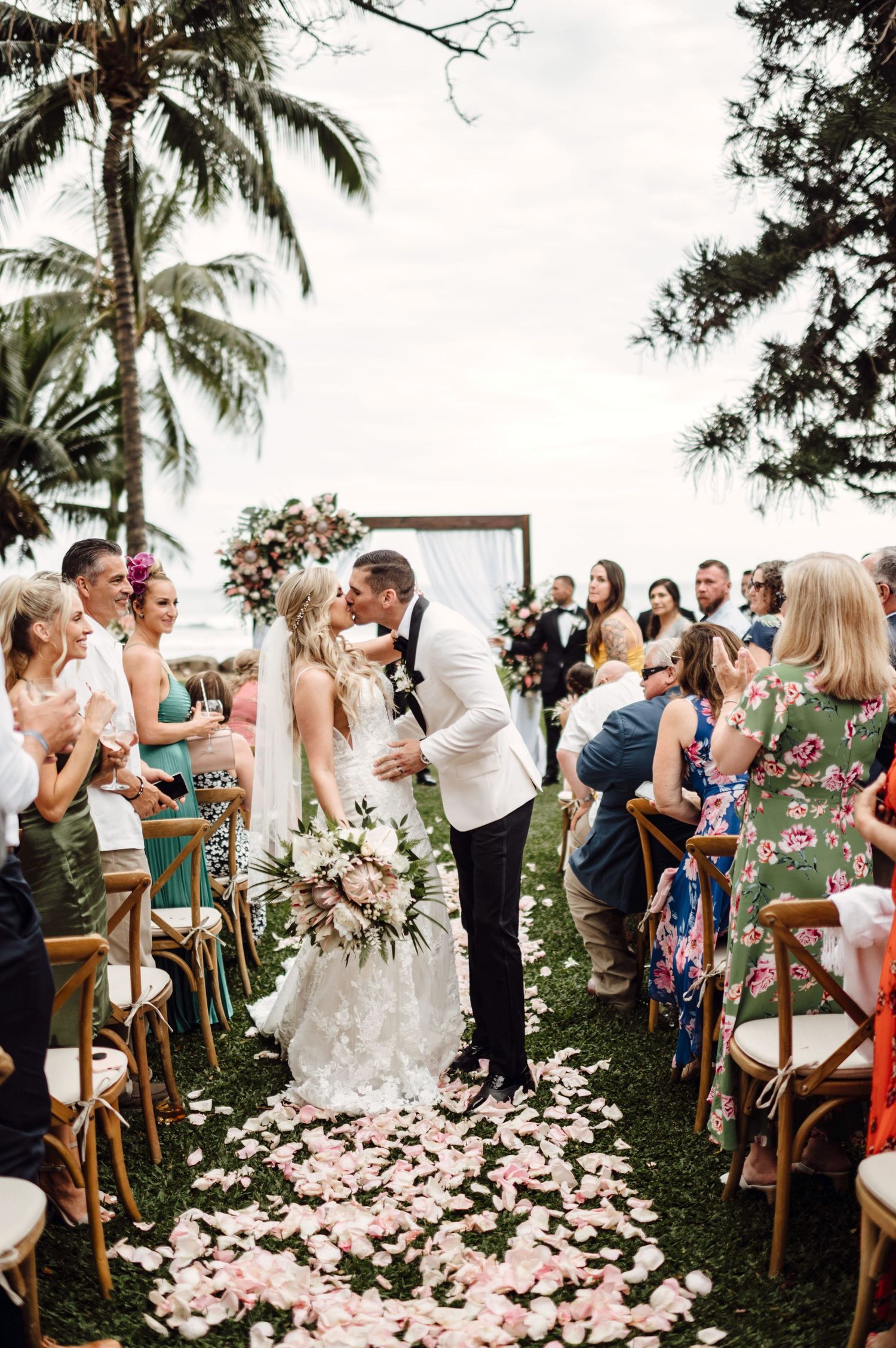 Bride and groom share a first kiss at outdoor wedding ceremony aisle lined with pink rose petals and tropical palm trees.