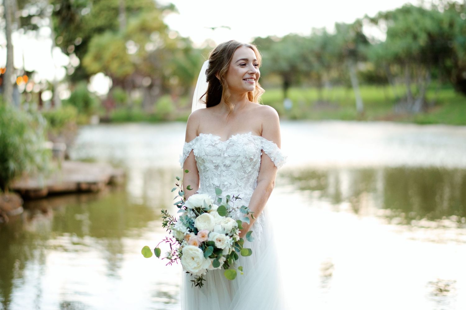 Smiling bride in off-shoulder lace gown holding white floral bouquet standing near serene lake outdoors.