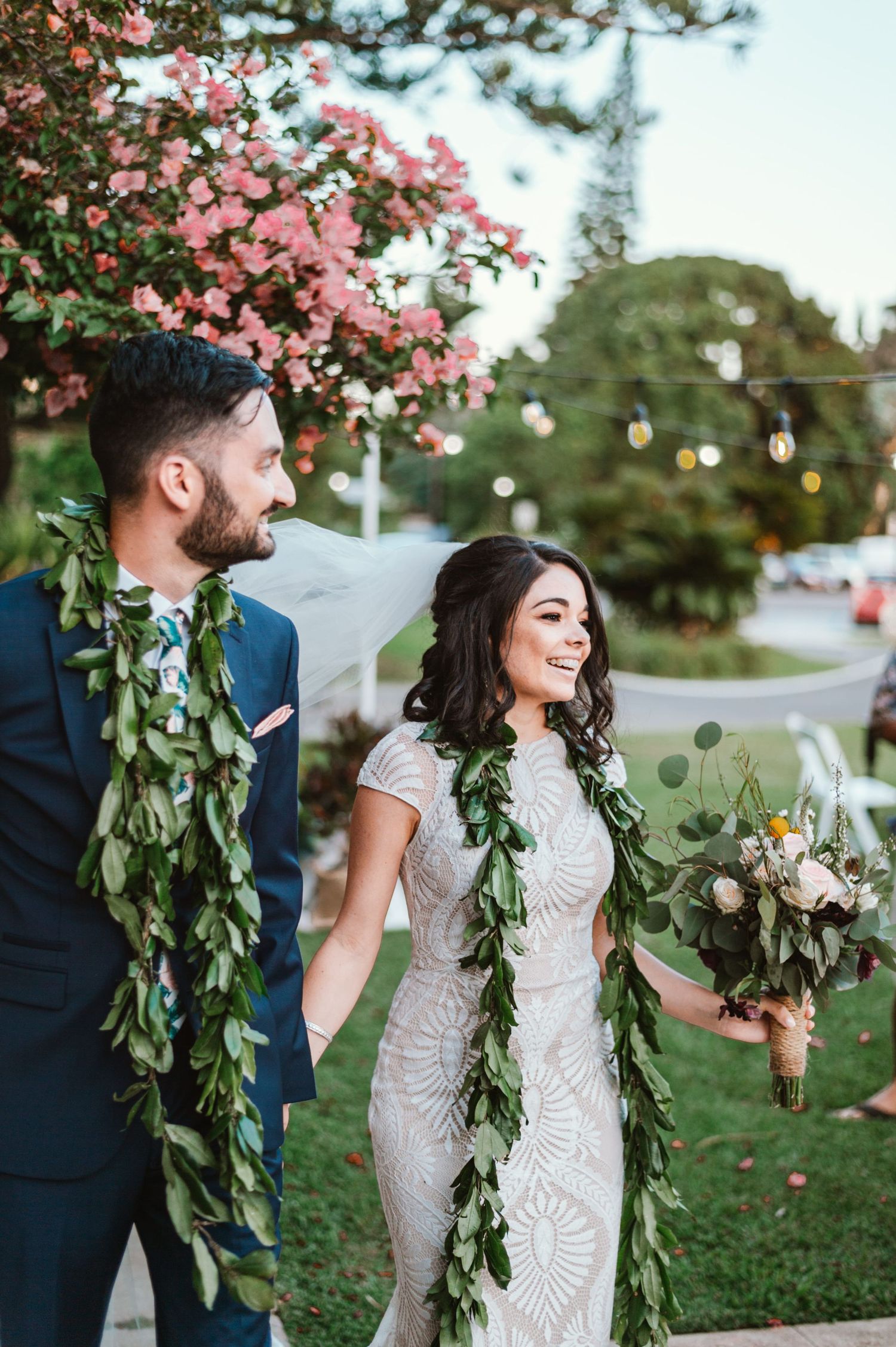 Bride and groom wearing traditional Hawaiian leaf leis, holding a bouquet, smiling outdoors near pink flowering trees.