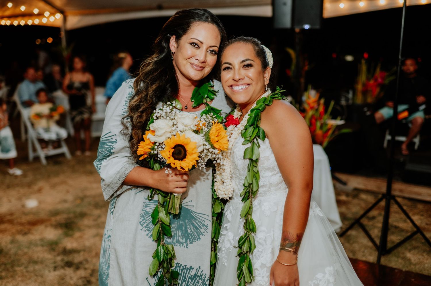 Bride and maid of honor wearing traditional Hawaiian leis share a joyful moment at their outdoor wedding reception.