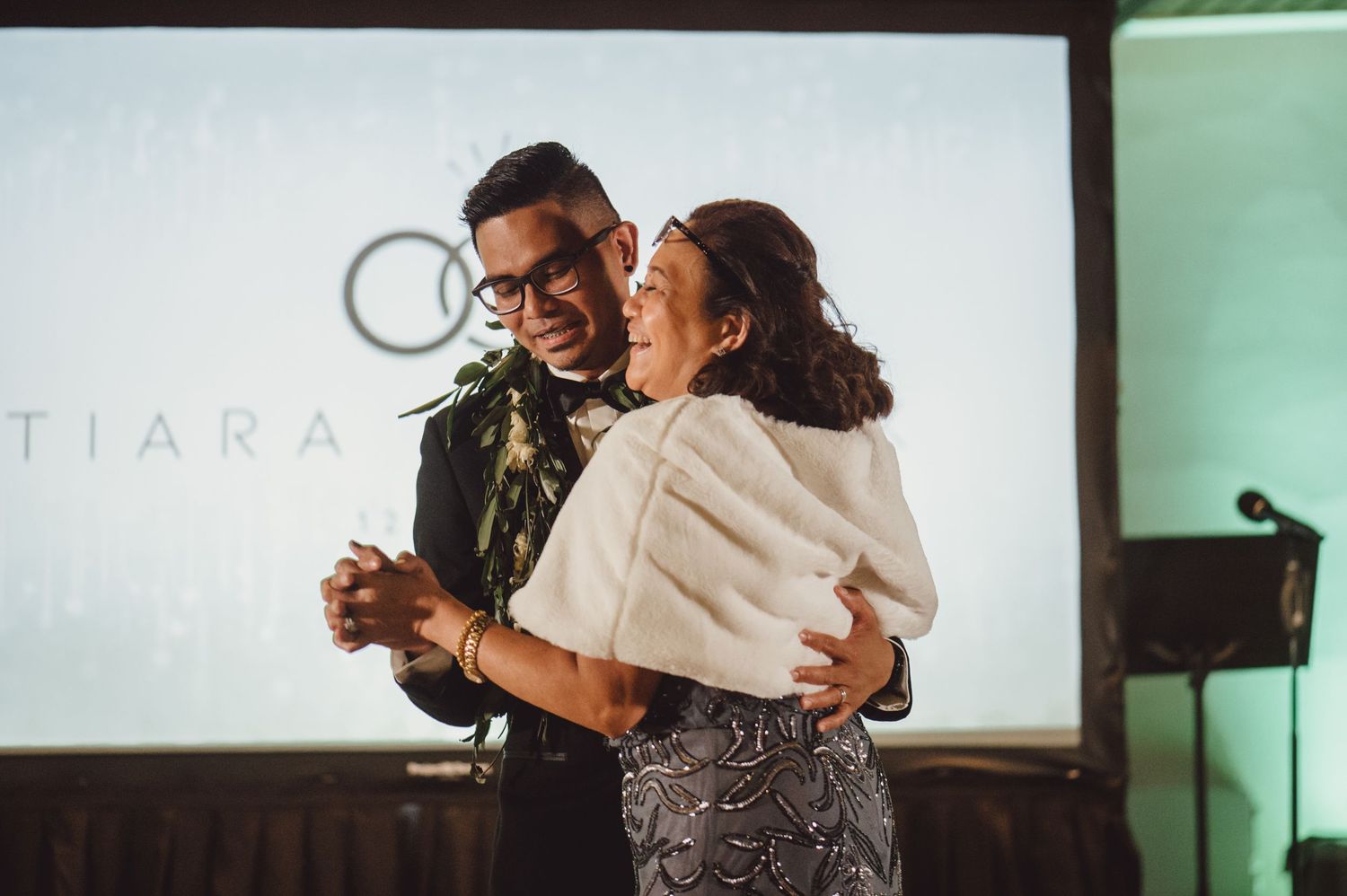 Couple dancing together at an indoor wedding reception with projection screen in background.
