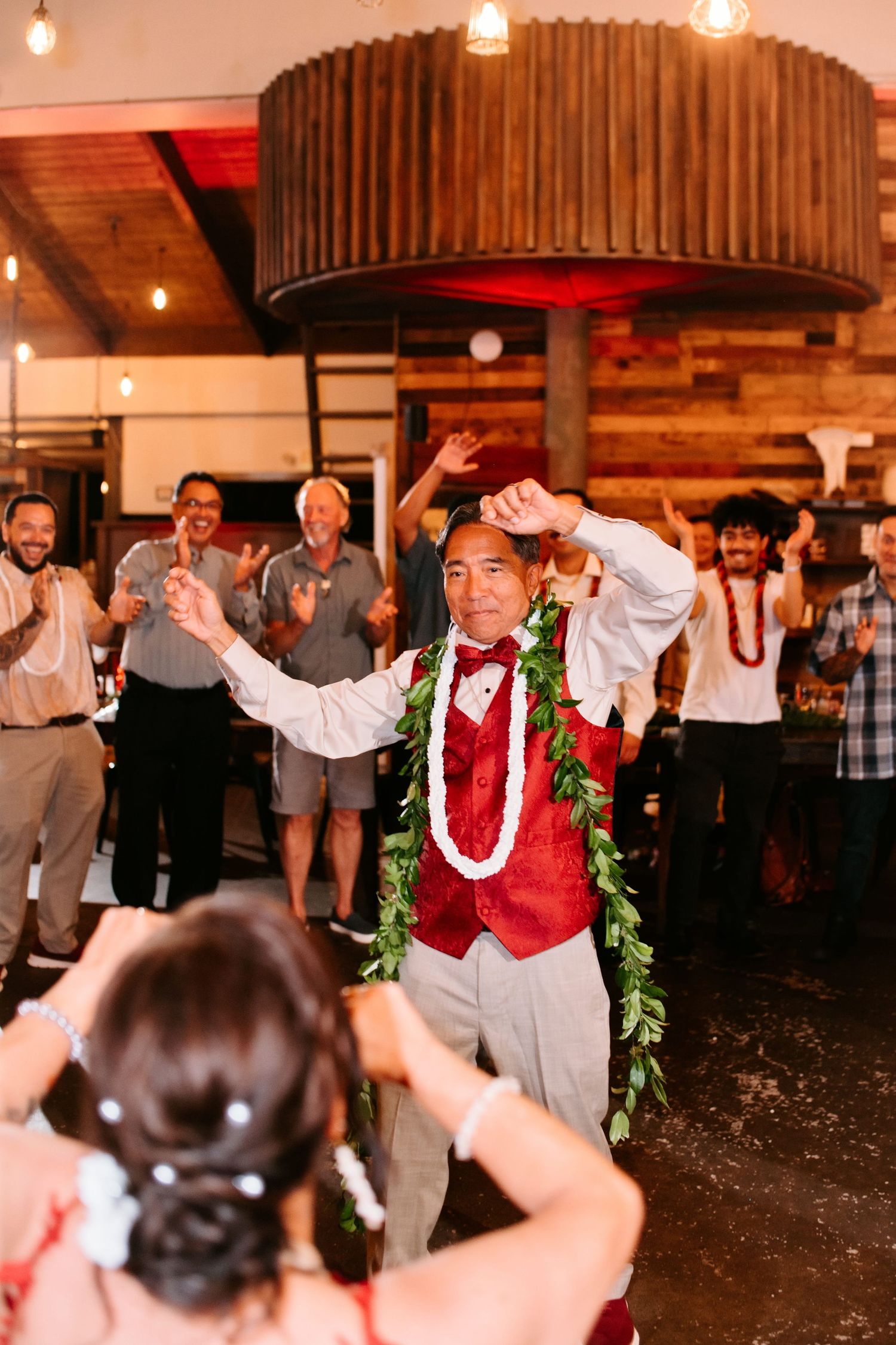 Groom dancing in red vest and green lei surrounded by cheering wedding guests at reception.