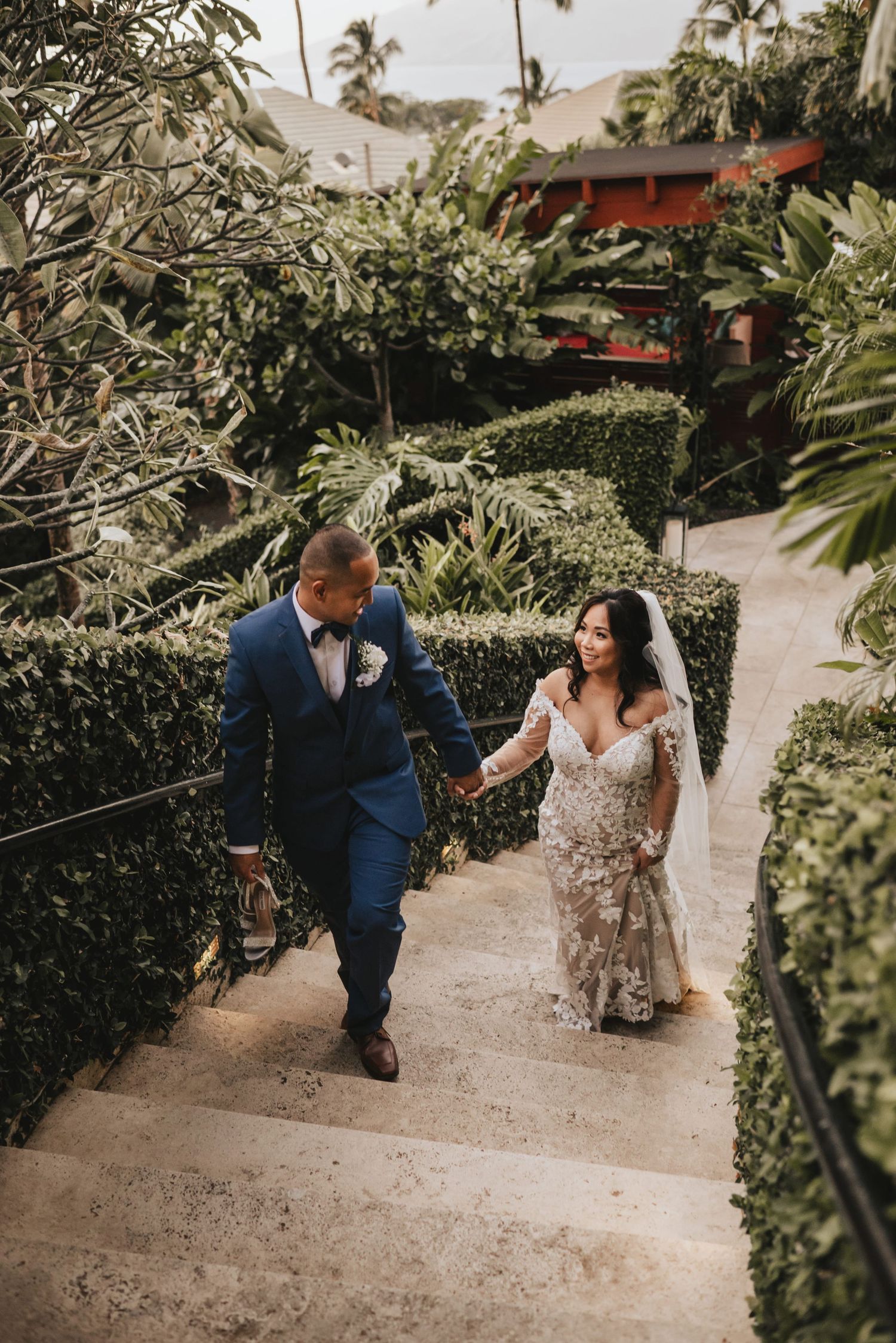 Aerial view of bride and groom walking hand-in-hand down tropical garden stairway on wedding day.