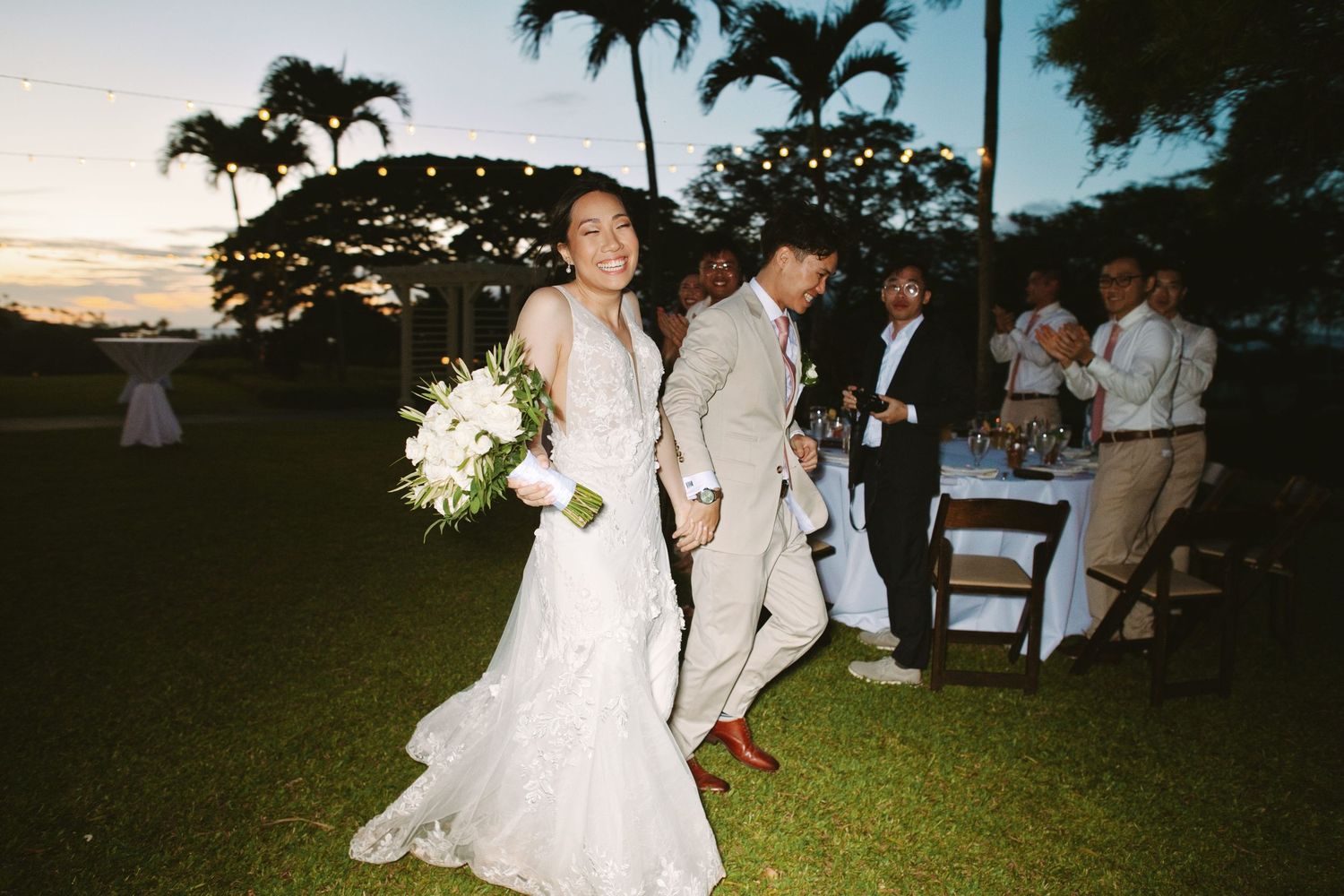 Bride in lace gown and groom in tan suit walk across lawn at outdoor wedding reception at sunset.