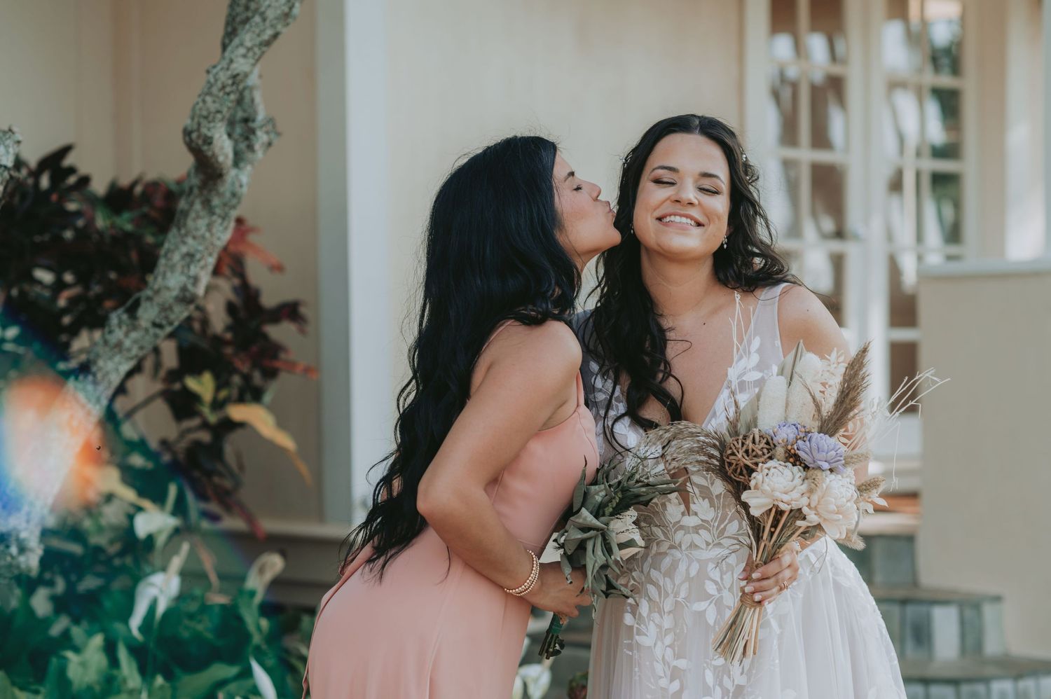 Bride in lace gown holds bouquet while bridesmaid in pink dress gives her a kiss on the cheek outdoors.