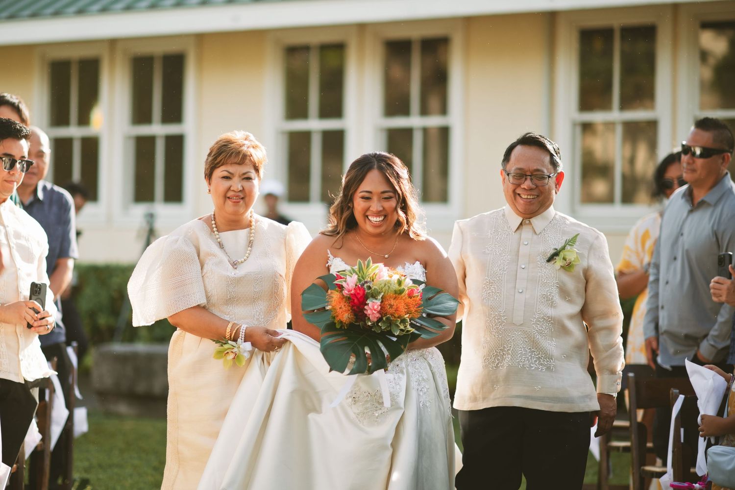 Bride walks down the aisle with parents during outdoor wedding ceremony, holding tropical bouquet.