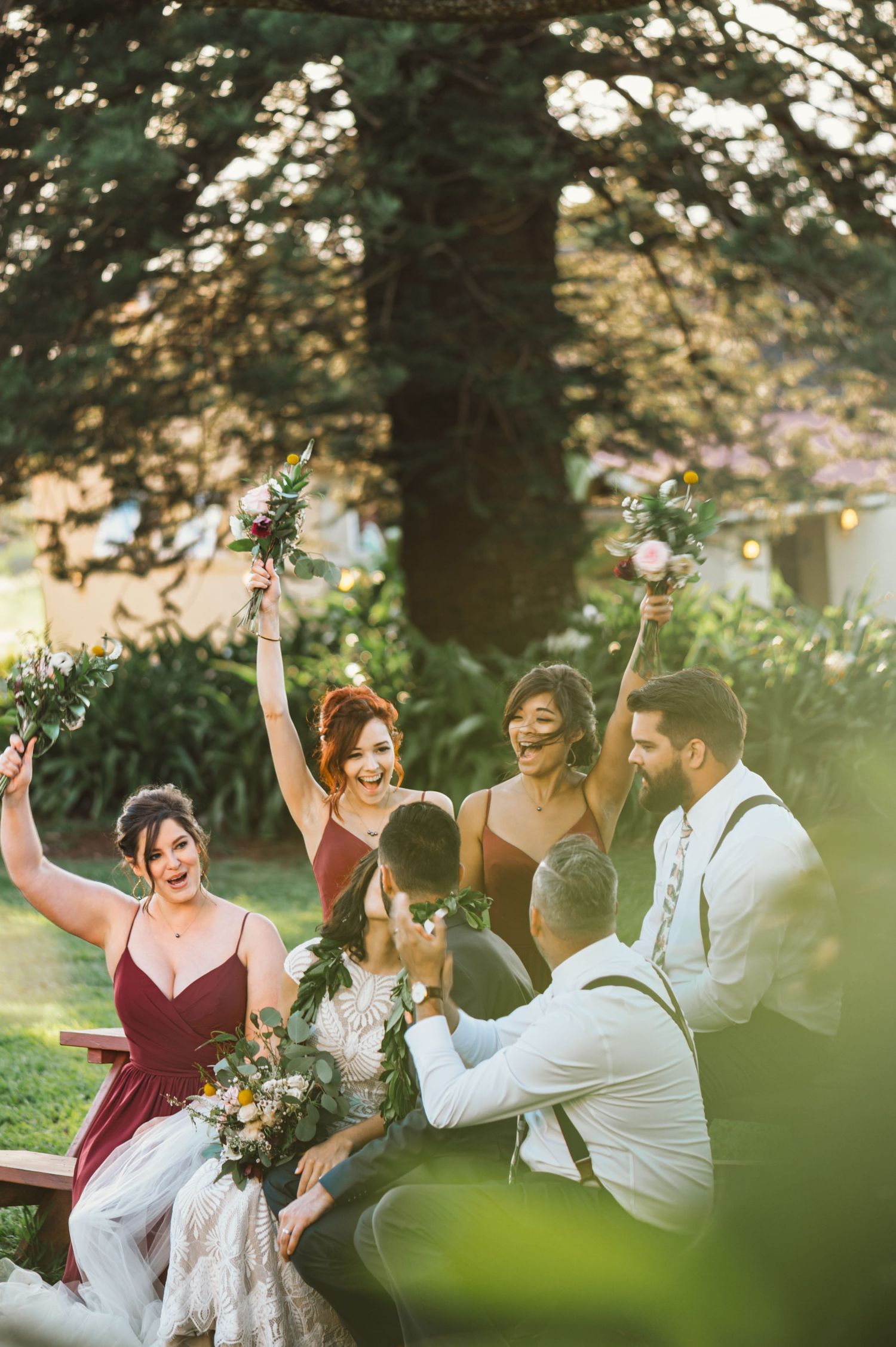 Joyful wedding party celebrating outdoors, bridesmaids in burgundy dresses raising bouquets among lush garden greenery.