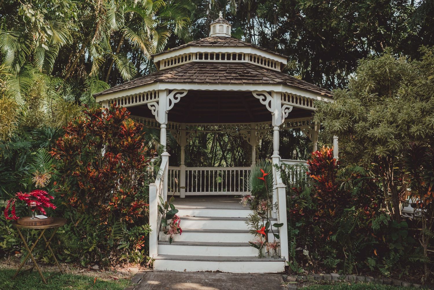 White gazebo with ornate trim surrounded by lush tropical plants and colorful flowers in a garden setting.