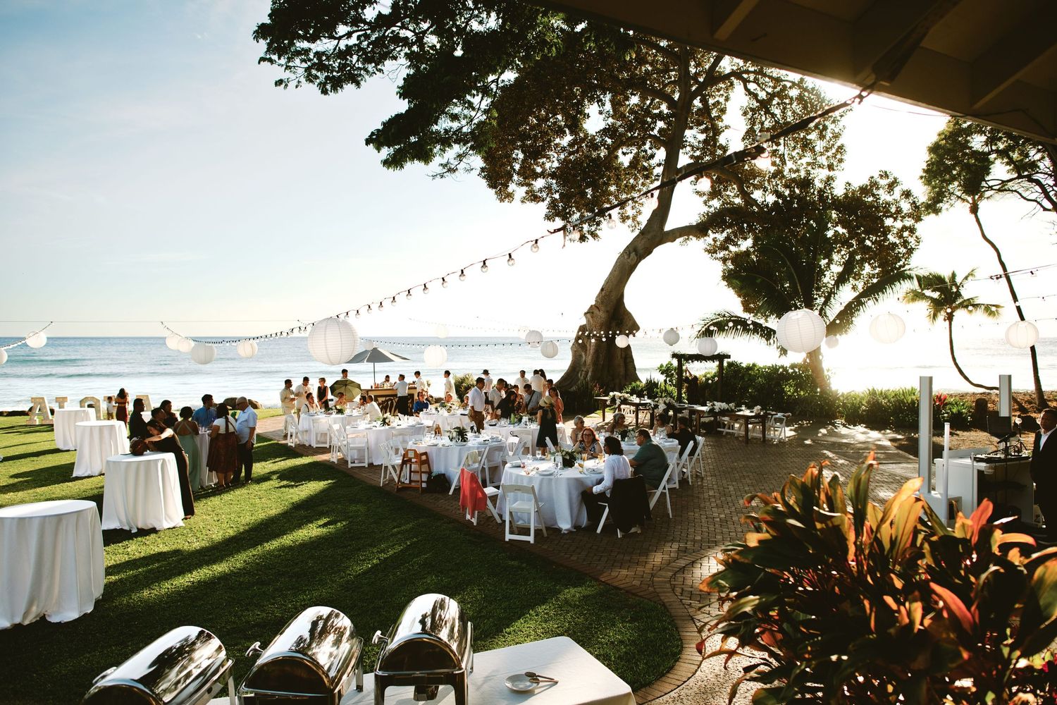 Elegant outdoor beachside wedding reception with white-draped tables, string lights, and tropical trees at sunset.