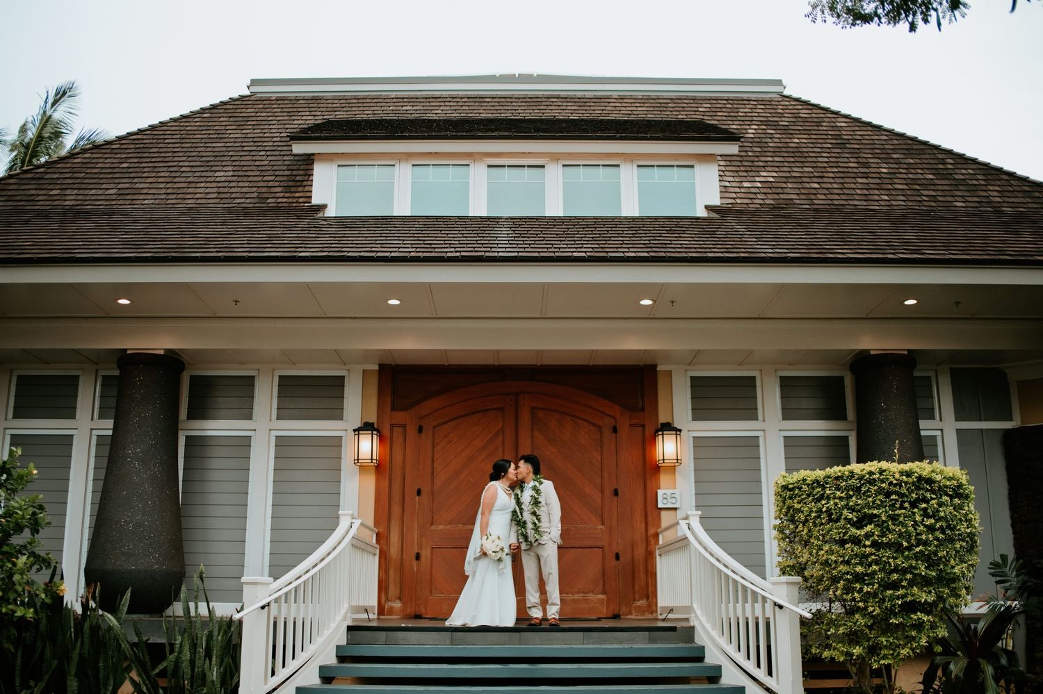 Bride and groom share a kiss on steps of elegant home with grand wooden doors on their wedding day.