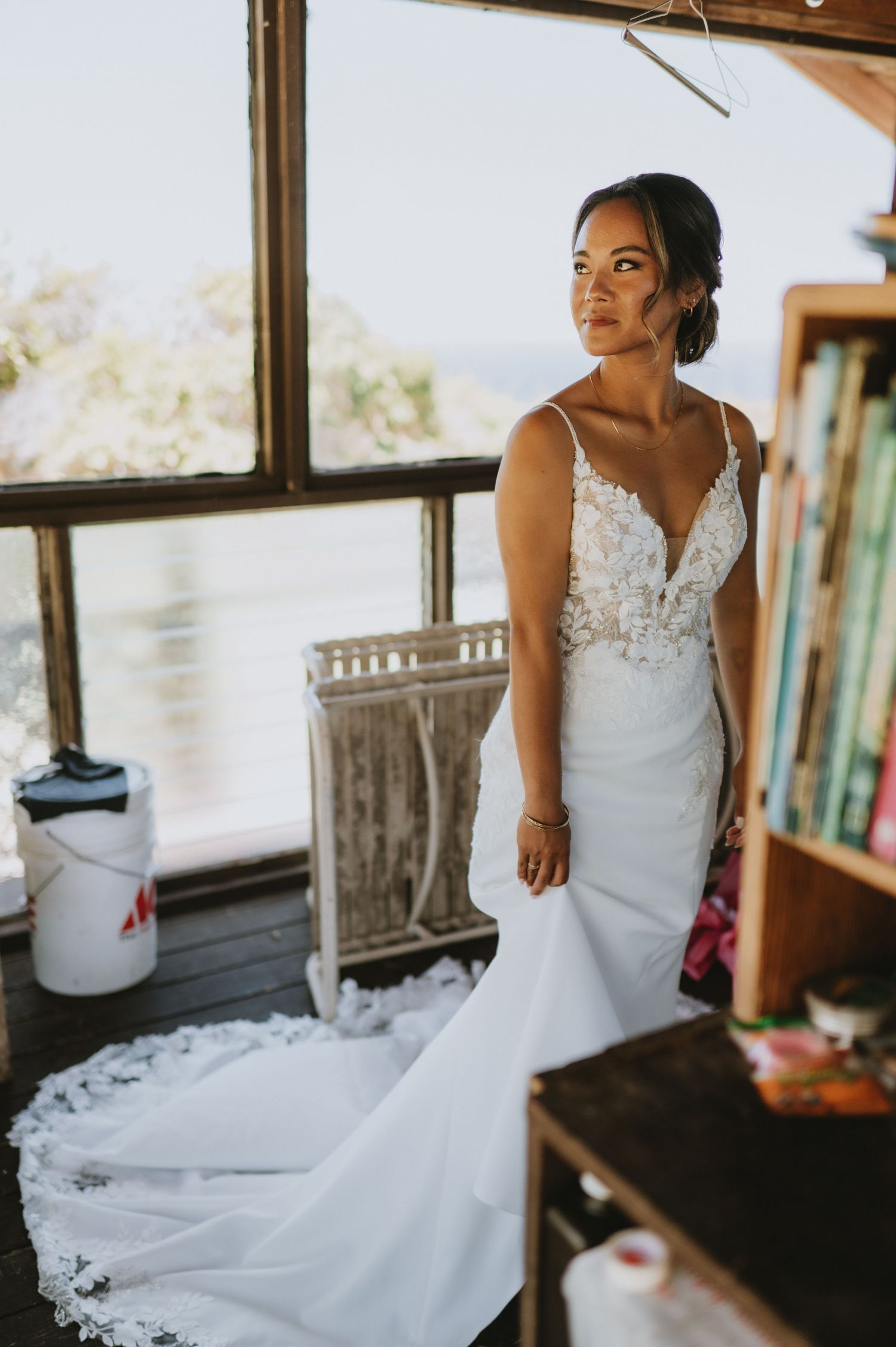 Bride in white lace mermaid wedding gown with train stands by rustic wooden window with ocean view.