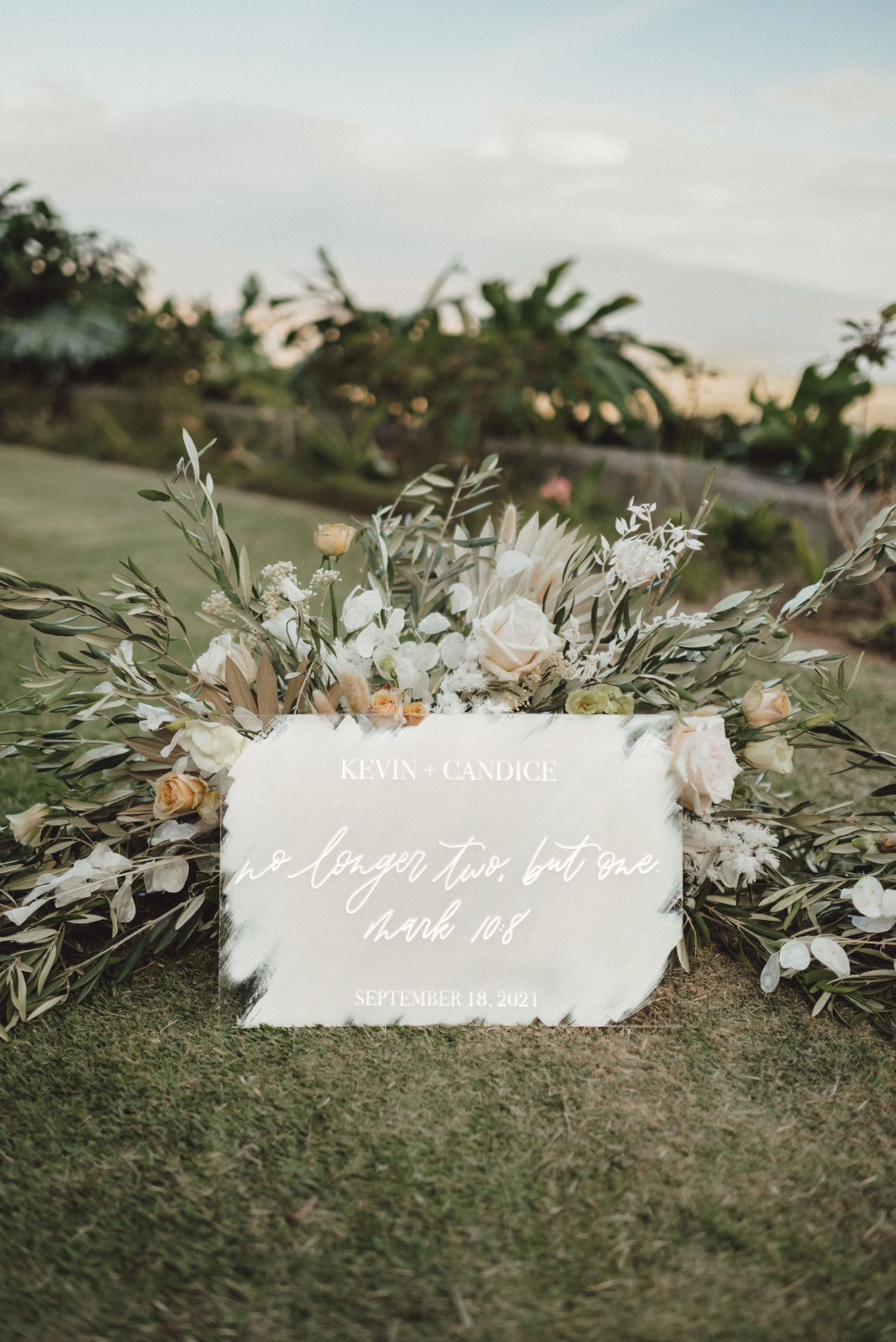 An elegant wedding sign surrounded by white floral arrangements and greenery against a tropical backdrop.