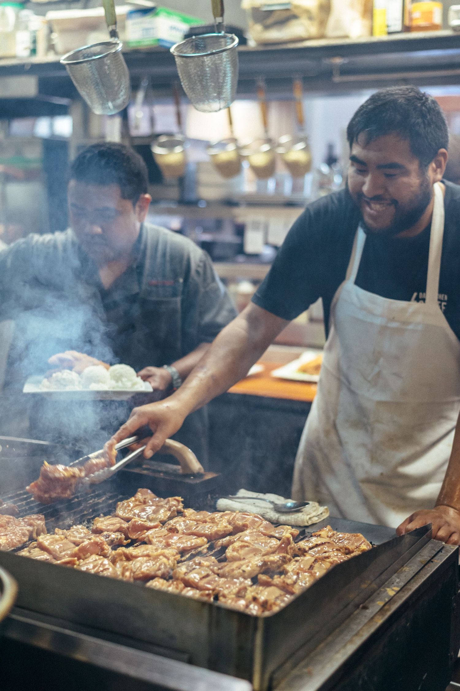 Cooks grilling meat on a large commercial flat top grill with smoke rising.