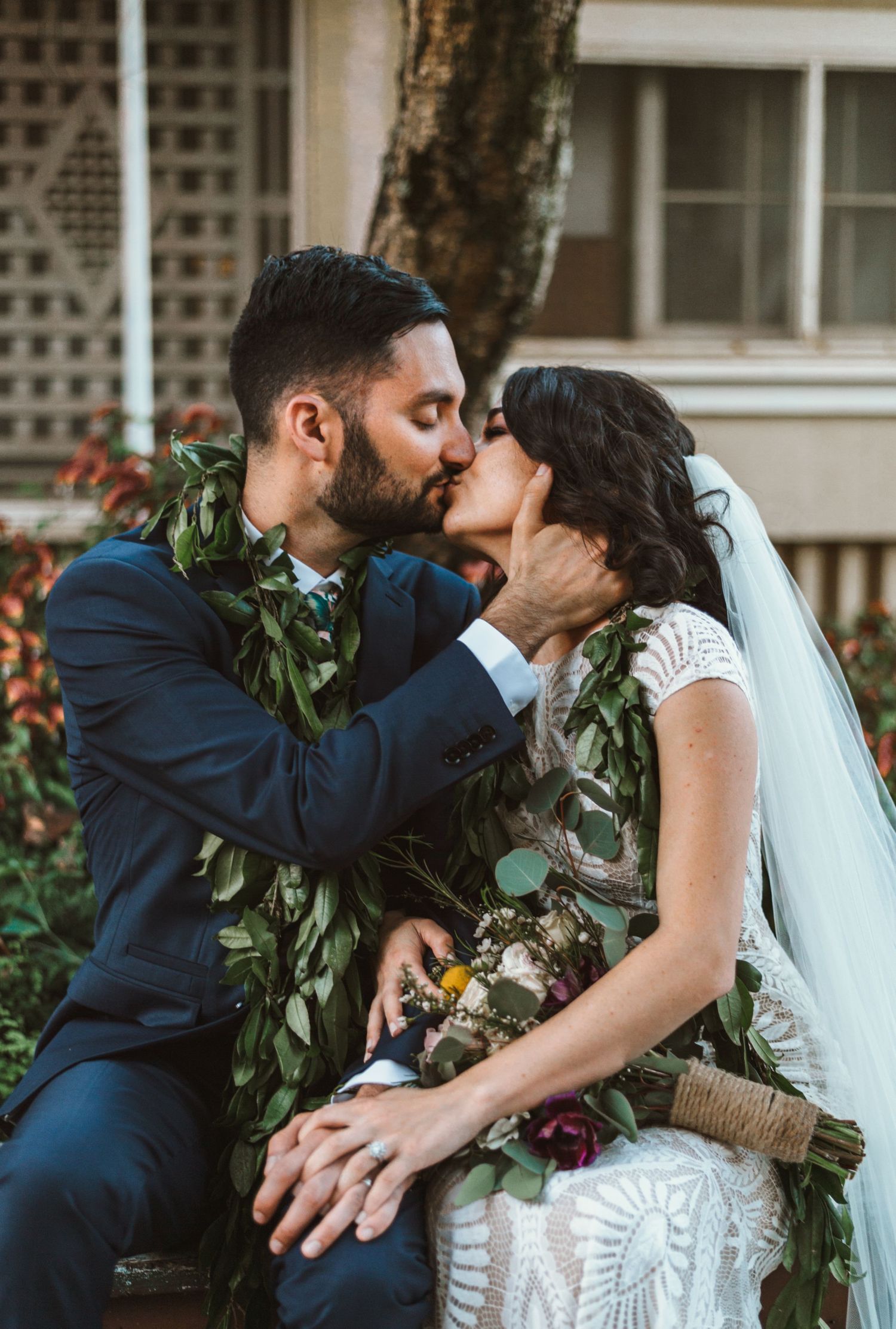 A romantic wedding kiss between newlyweds wearing traditional Hawaiian leis and formal attire in an outdoor garden setting.