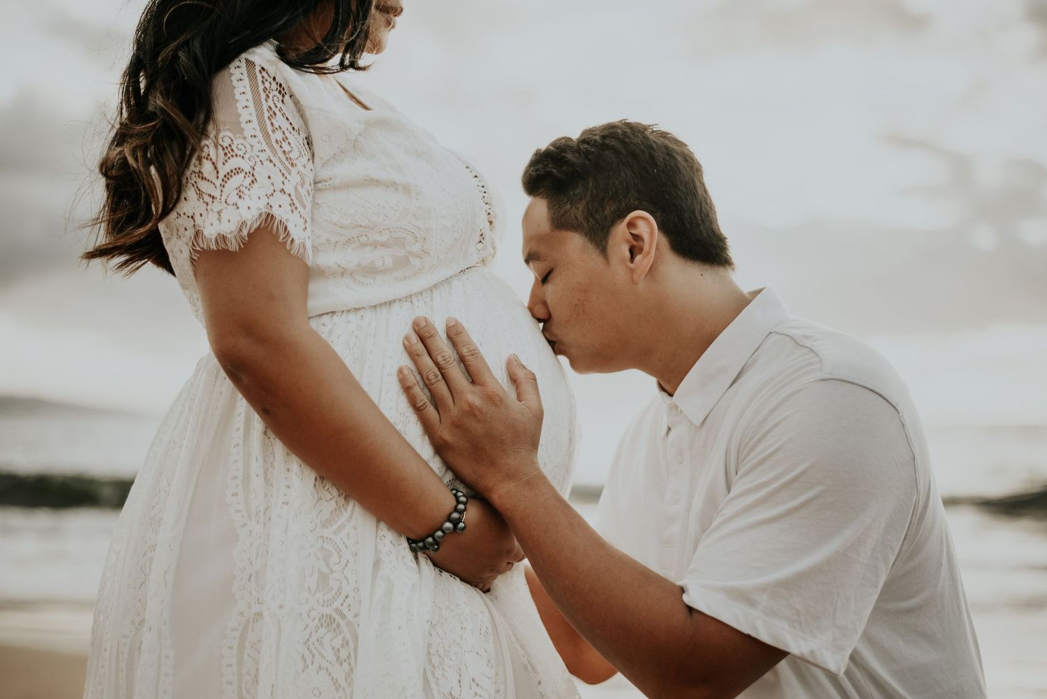 Intimate maternity moment captured on rocky beach with white lace dress.