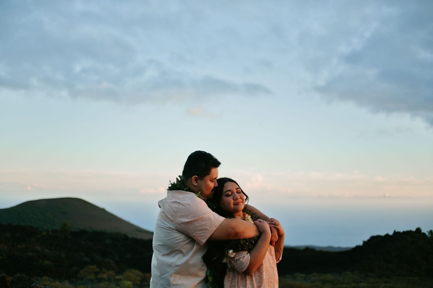 Couple embracing at sunset on a Hawaiian hilltop, wearing lei garlands with scenic green mountains behind them.
