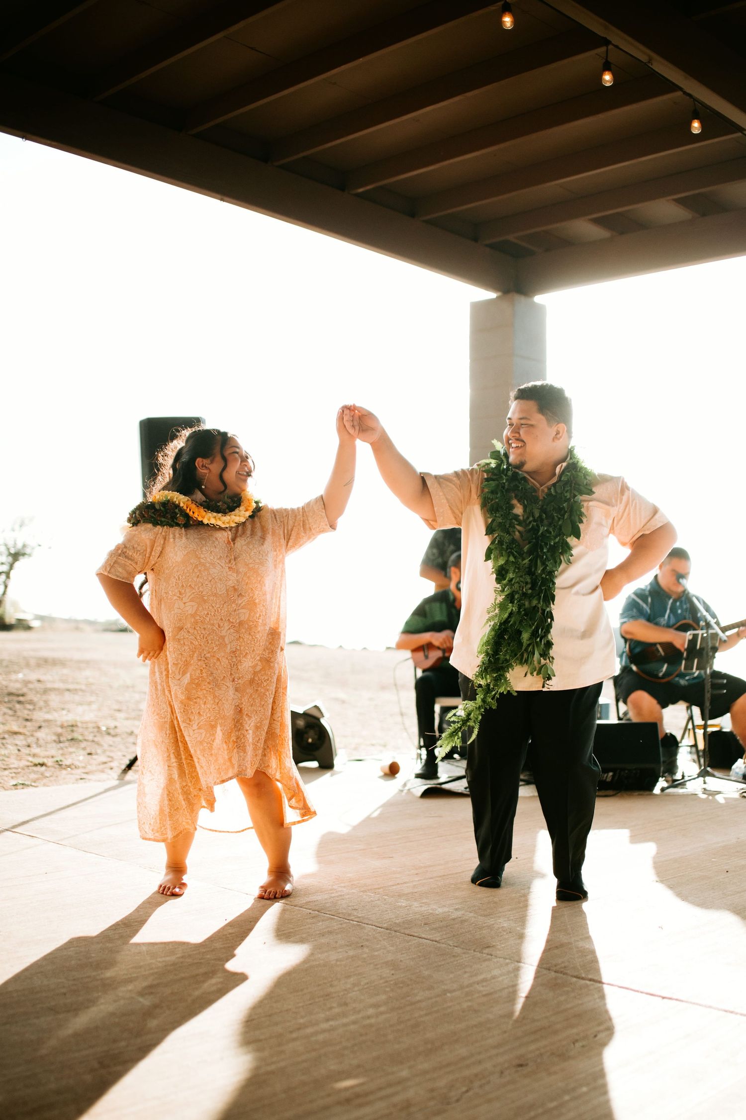 Couple dancing in leis at an outdoor Hawaiian celebration with live music and warm sunset lighting.