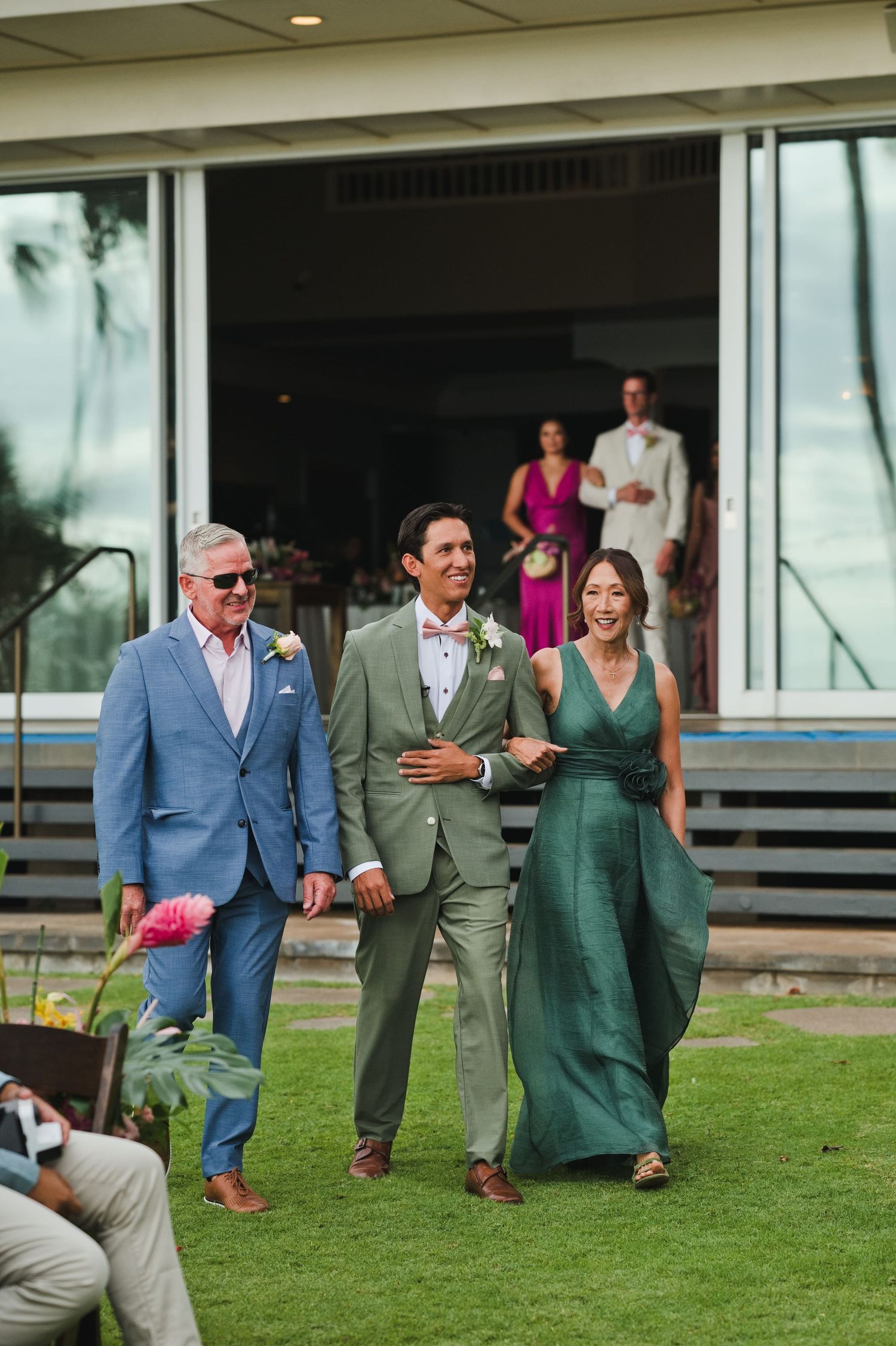 Groom in sage green suit walks down the aisle flanked by smiling family members at an outdoor wedding.