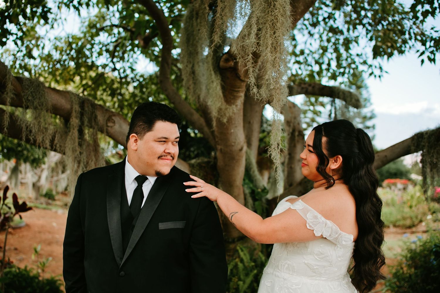 Bride in off-shoulder gown touches groom in black tuxedo during first look beneath a large tree in a nearby garden.