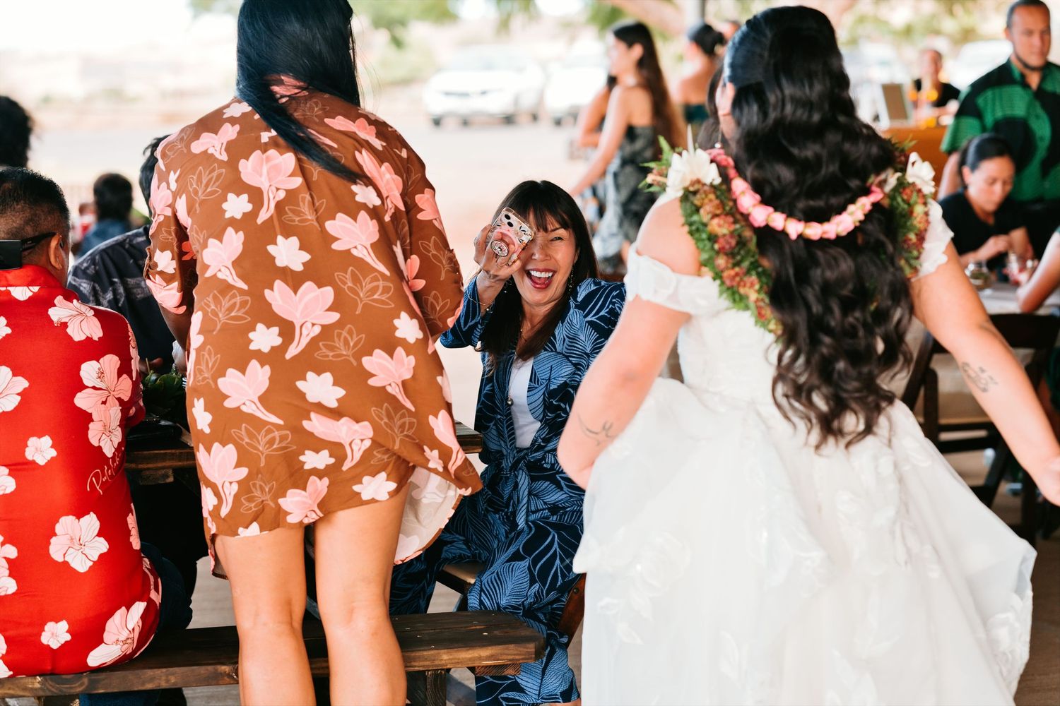 A bride in a white off-shoulder gown with a floral lei greets guests at an outdoor wedding reception.