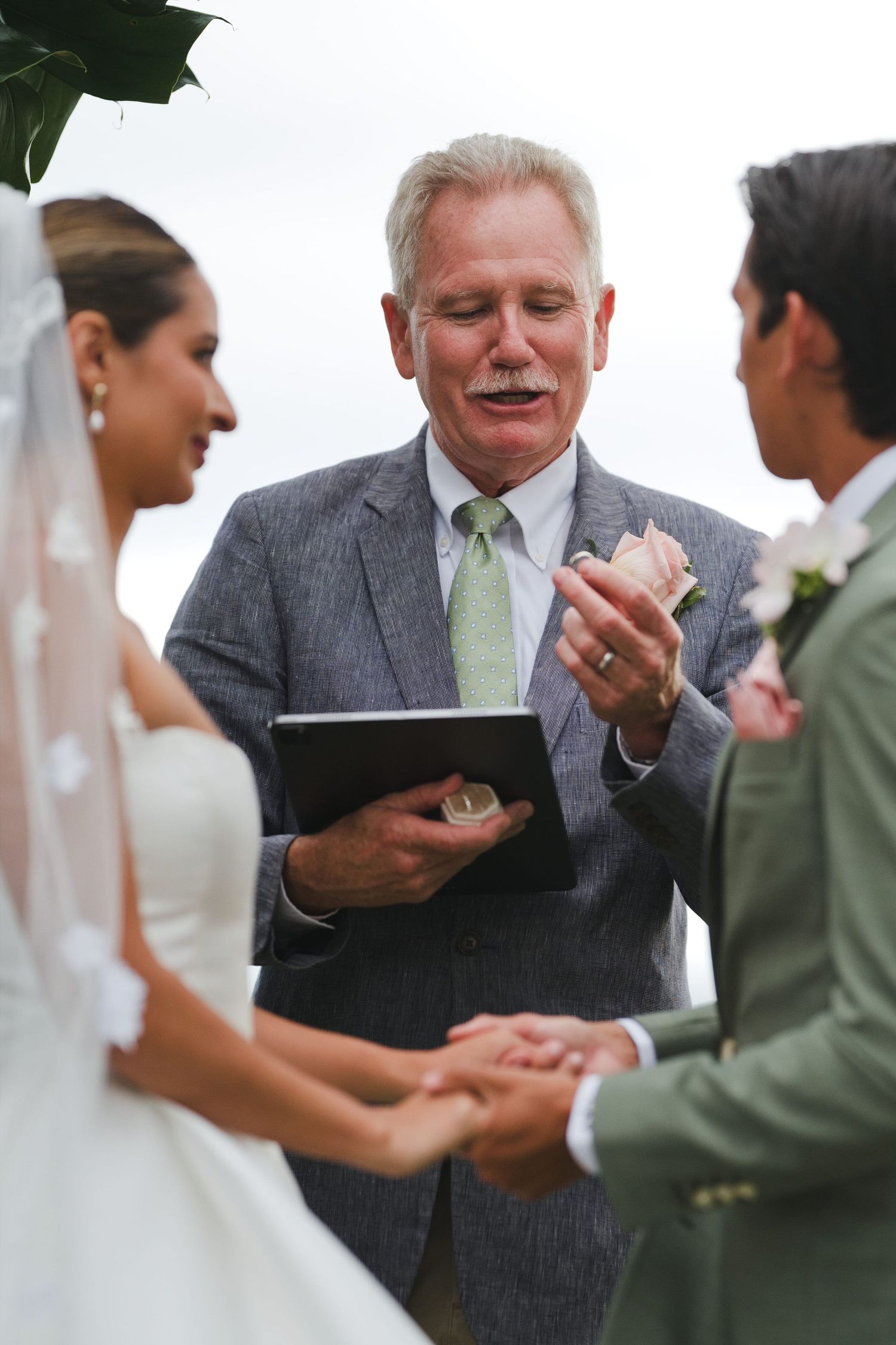 Officiant in gray suit reads vows from tablet at outdoor wedding ceremony as bride and groom hold hands.