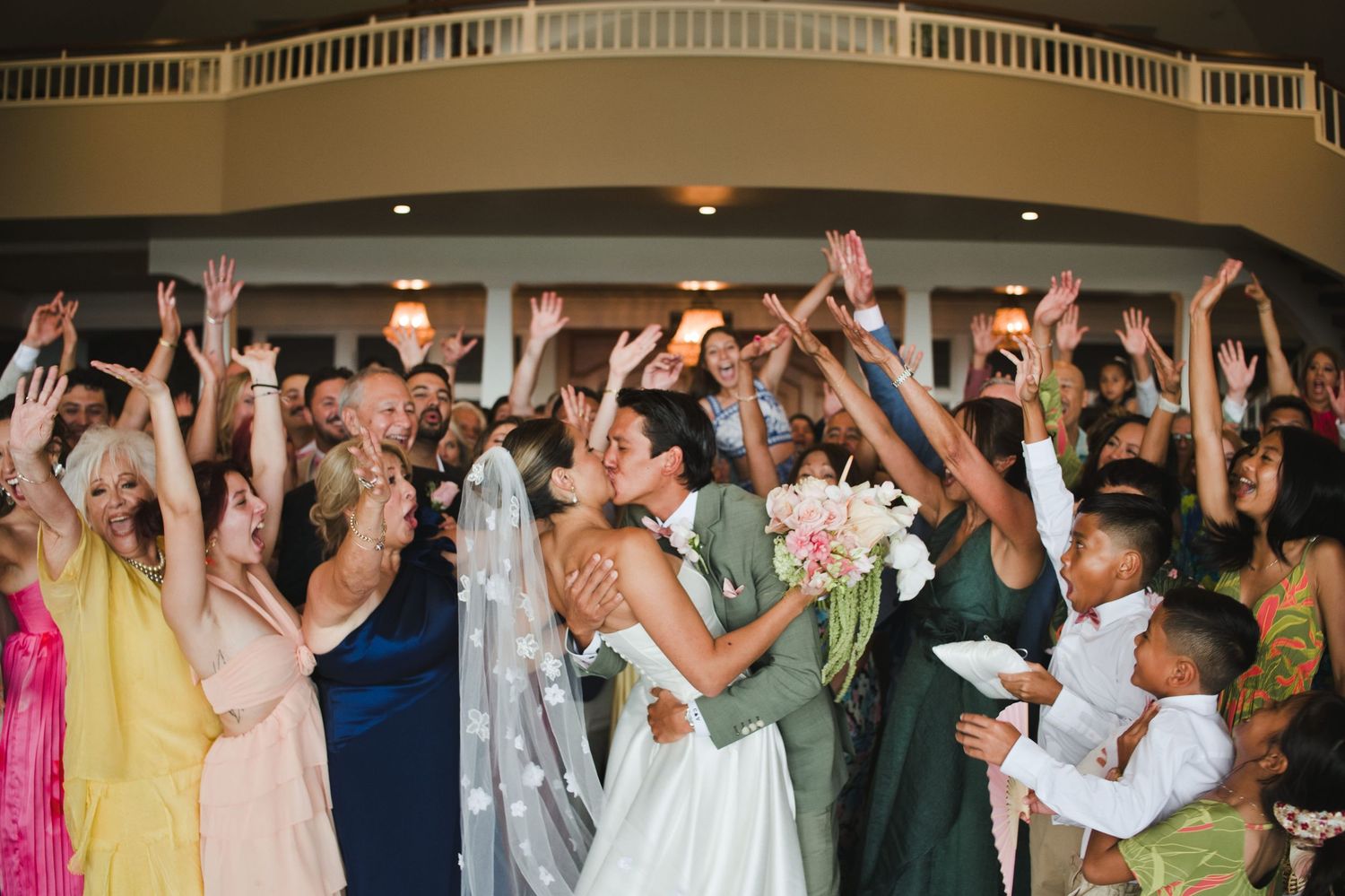 Bride and groom share a kiss surrounded by cheering wedding guests with raised arms at a festive indoor reception.