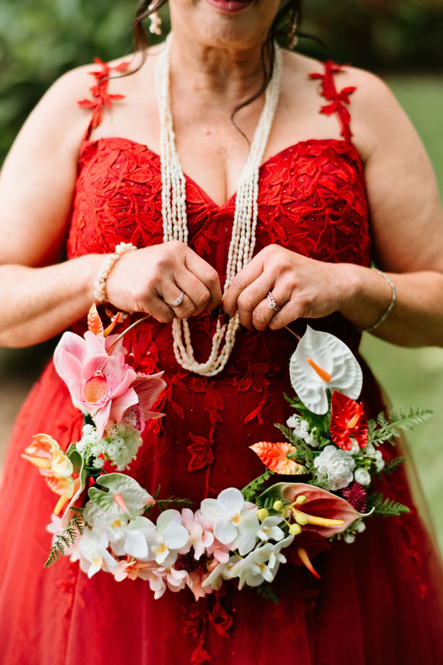 Woman in red lace dress holding a tropical floral wreath with orchids and anthuriums, wearing pearl necklaces.