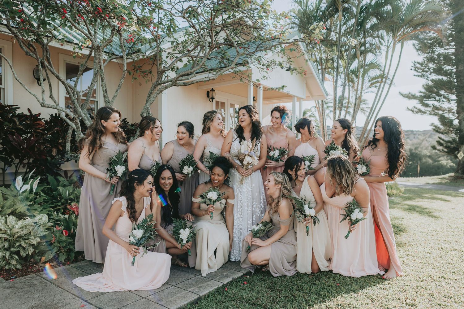Bride and bridesmaids in mismatched neutral gowns holding white bouquets gathered outside a tropical venue.