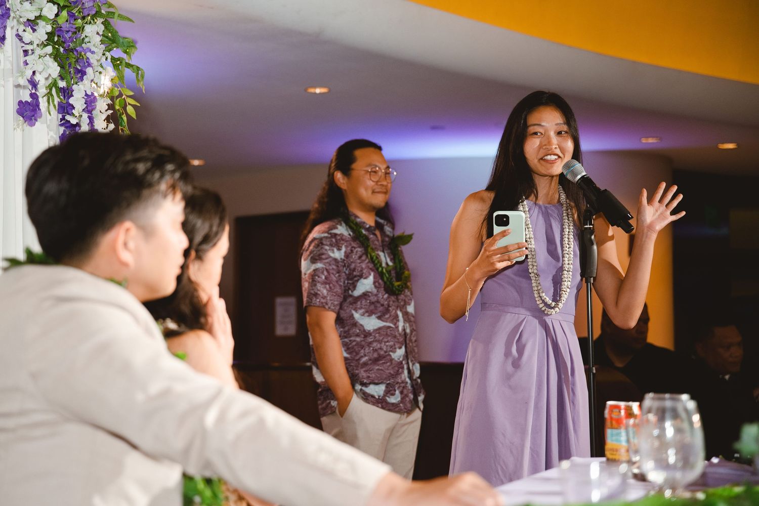 Woman in purple dress giving a speech at a Hawaiian wedding reception while guests listen attentively.
