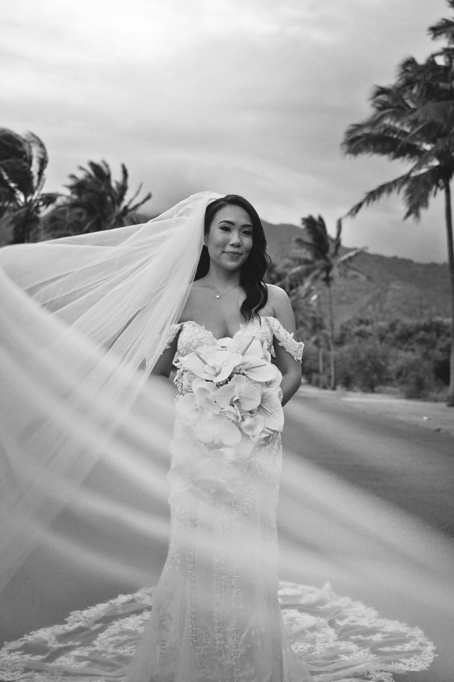 Black and white photo of a bride in lace gown with flowing veil, holding bouquet, standing near palm trees and mountains.